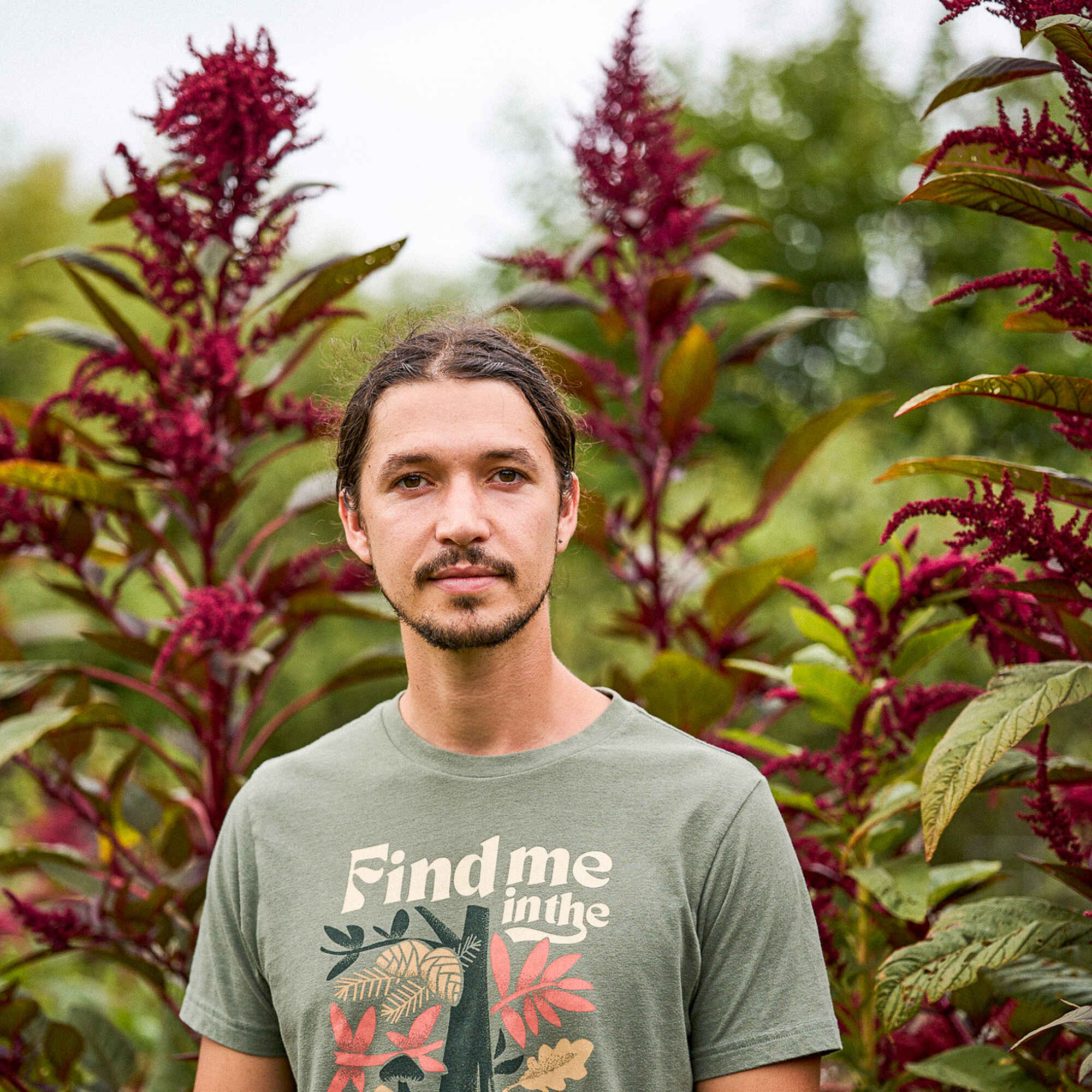 Bohdi Vasilopoulos standing in front of burgundy celosia flowers wearing a forest green t-shirt with nature-themed graphics