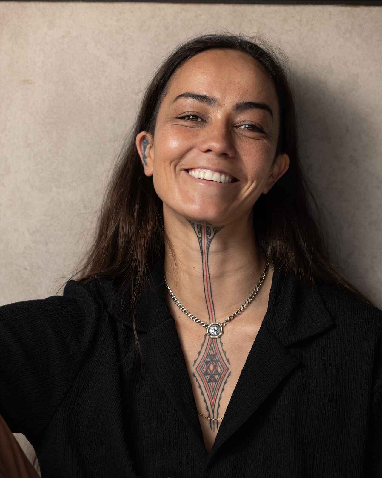 Briar Rose, a person with long dark hair and chest tattoos, smiling at the camera while wearing a black blazer and chain necklace