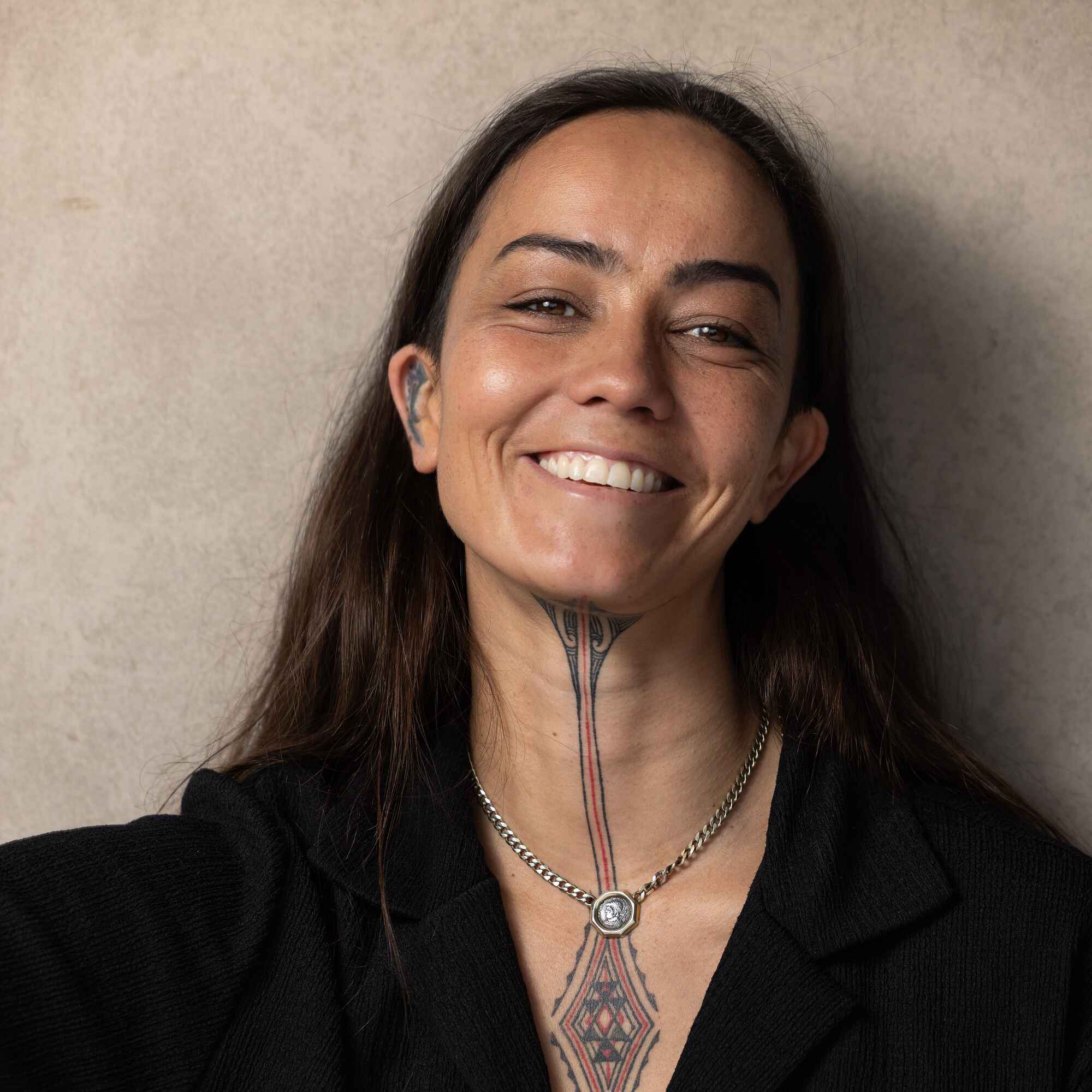 Briar Rose, a person with long dark hair and chest tattoos, smiling at the camera while wearing a black blazer and chain necklace