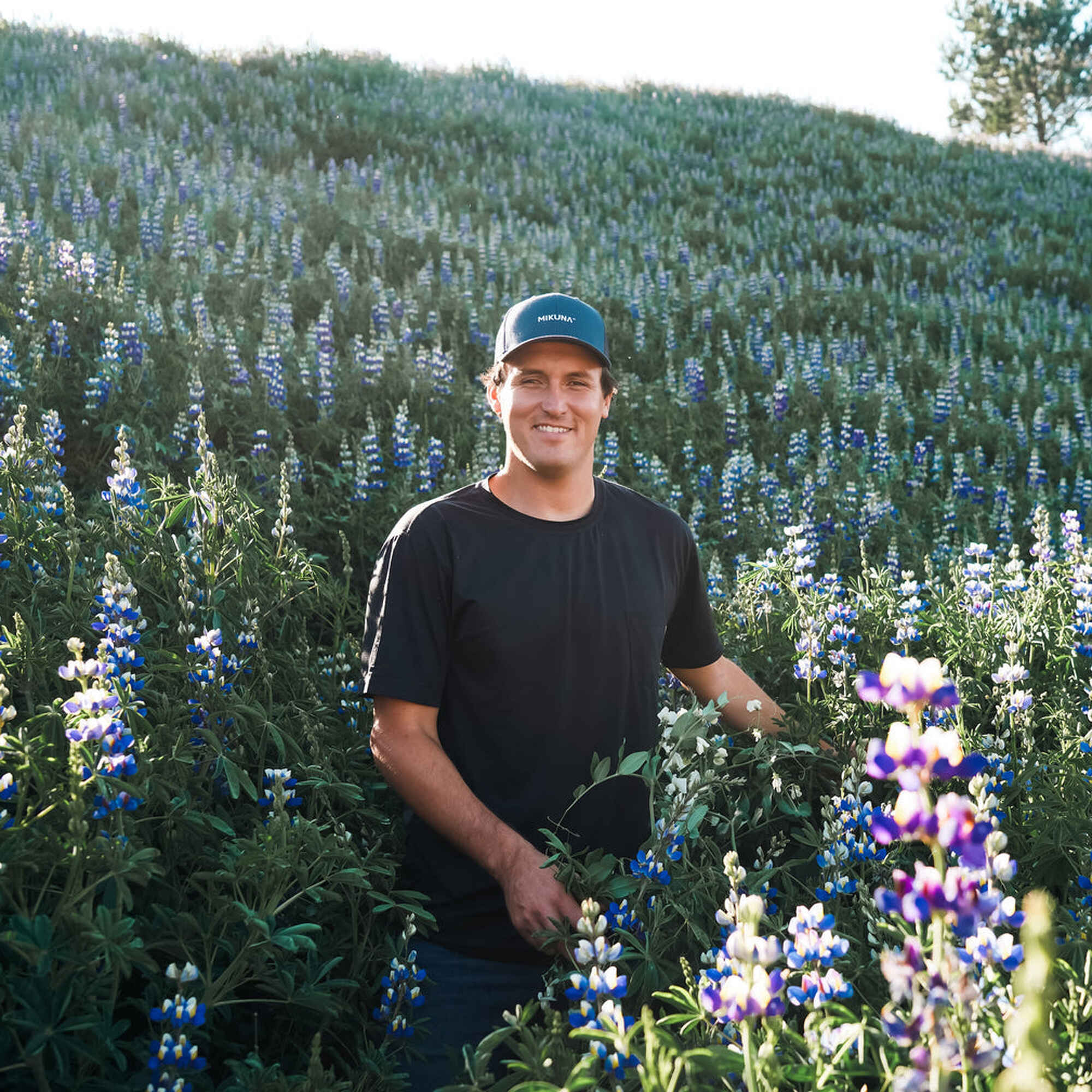 Ricky Echanique standing in a field of blooming lupine flowers with forested hills in the background