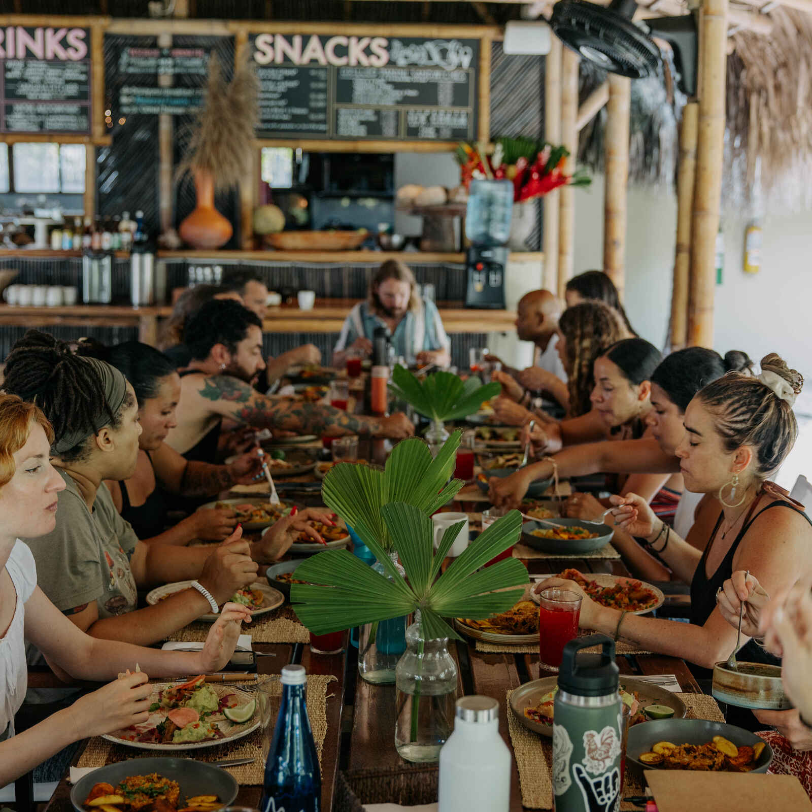 A diverse group of people sharing a communal meal at a long wooden table in a rustic open-air dining space