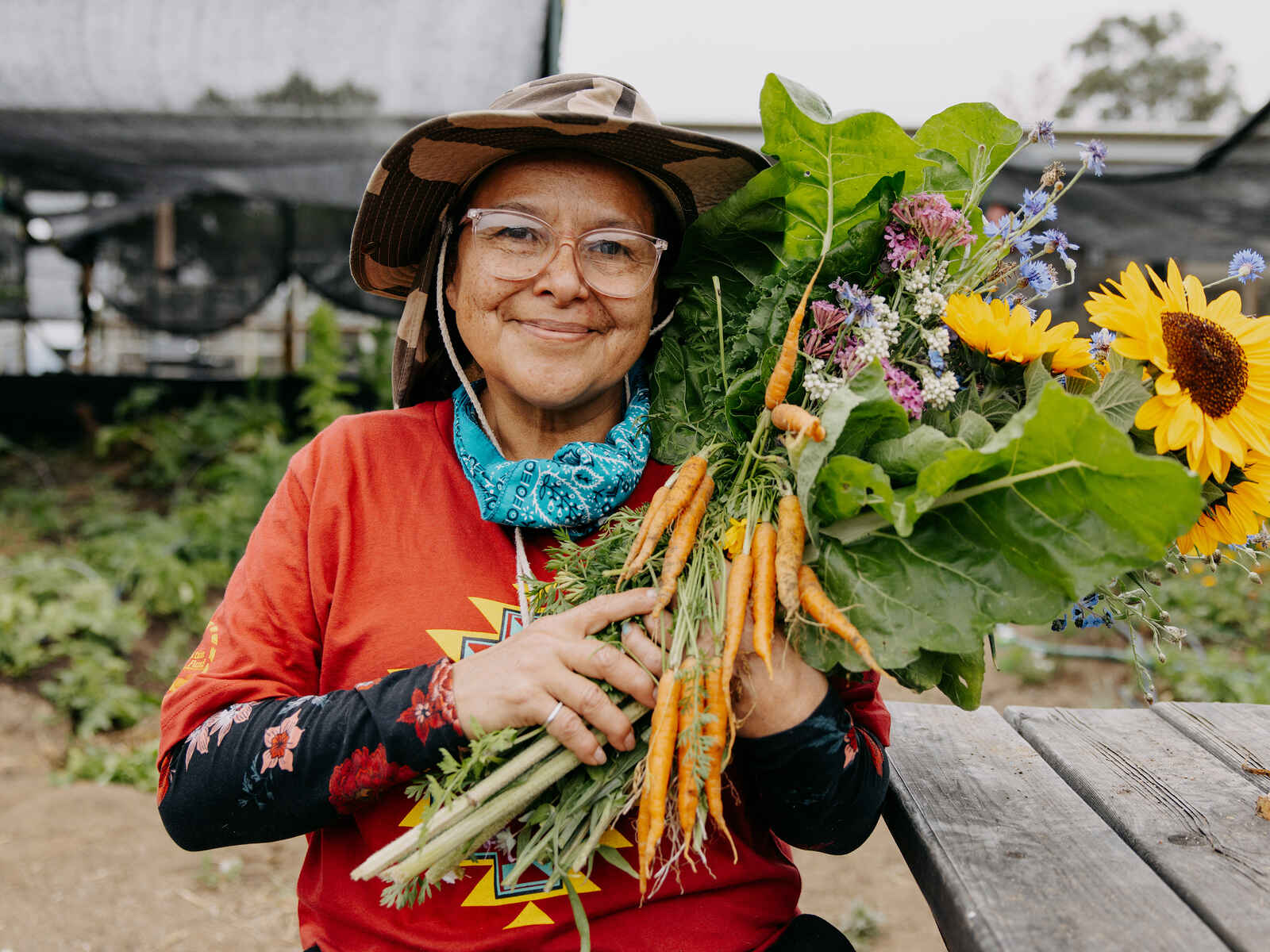 A farmer holds a fresh harvest of carrots, flowers, and vegetables in a greenhouse