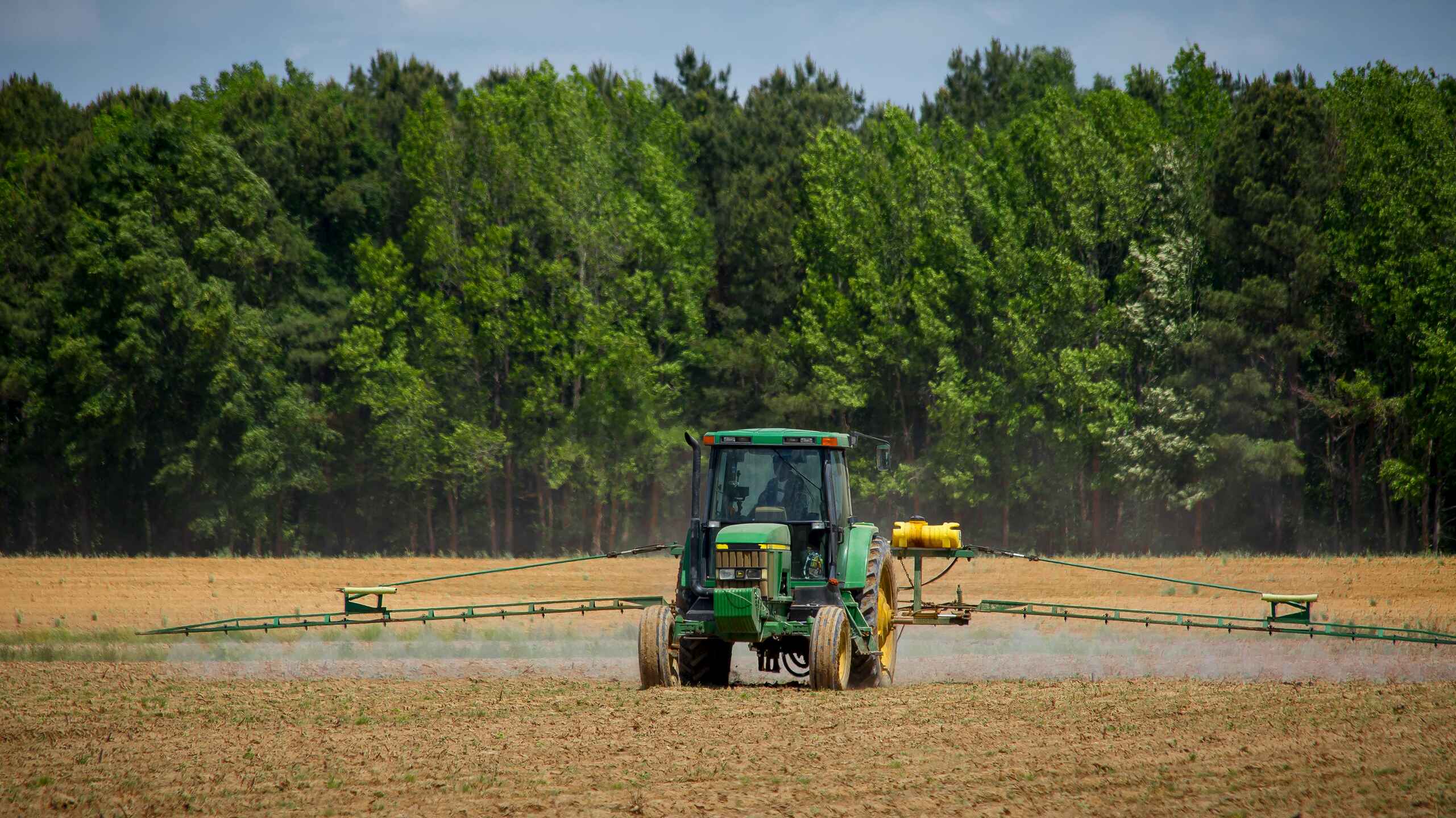 Green tractor with spray boom applying herbicide to a bare field with forest in background