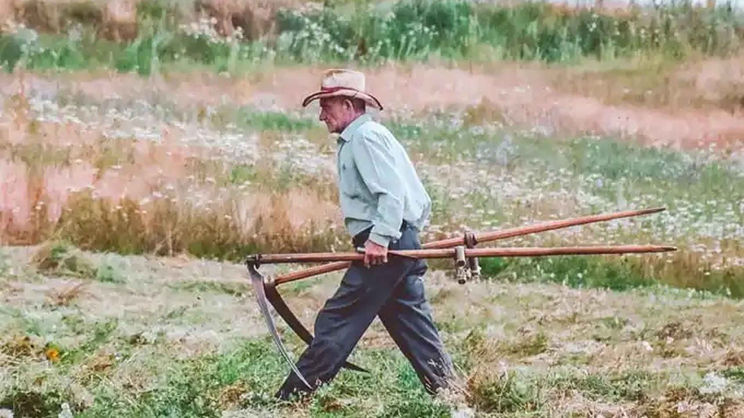 Farmer carrying a large plow across a field of exposed soil and sparse vegetation