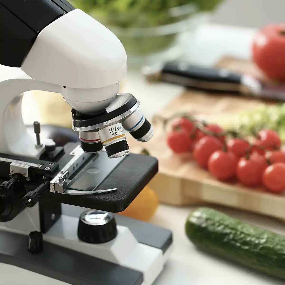 Microscope positioned over fresh produce including tomatoes and cucumber, symbolizing scientific examination of food quality and safety