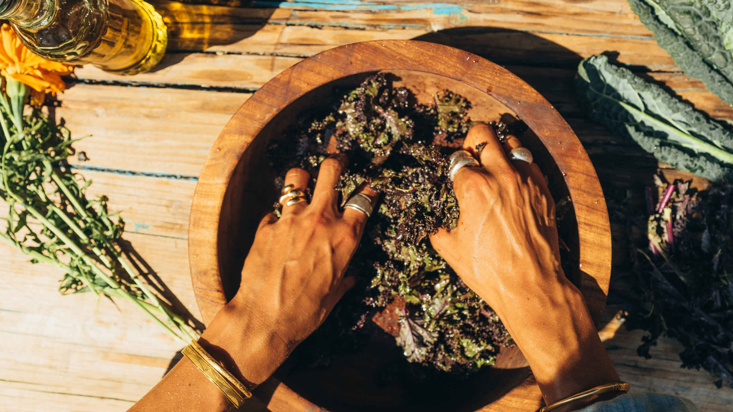 Hands massaging purple kale in a large wooden bowl on a sunlit plank table