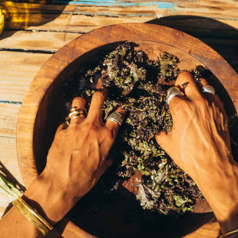 Hands massaging purple kale in a large wooden bowl on a sunlit plank table