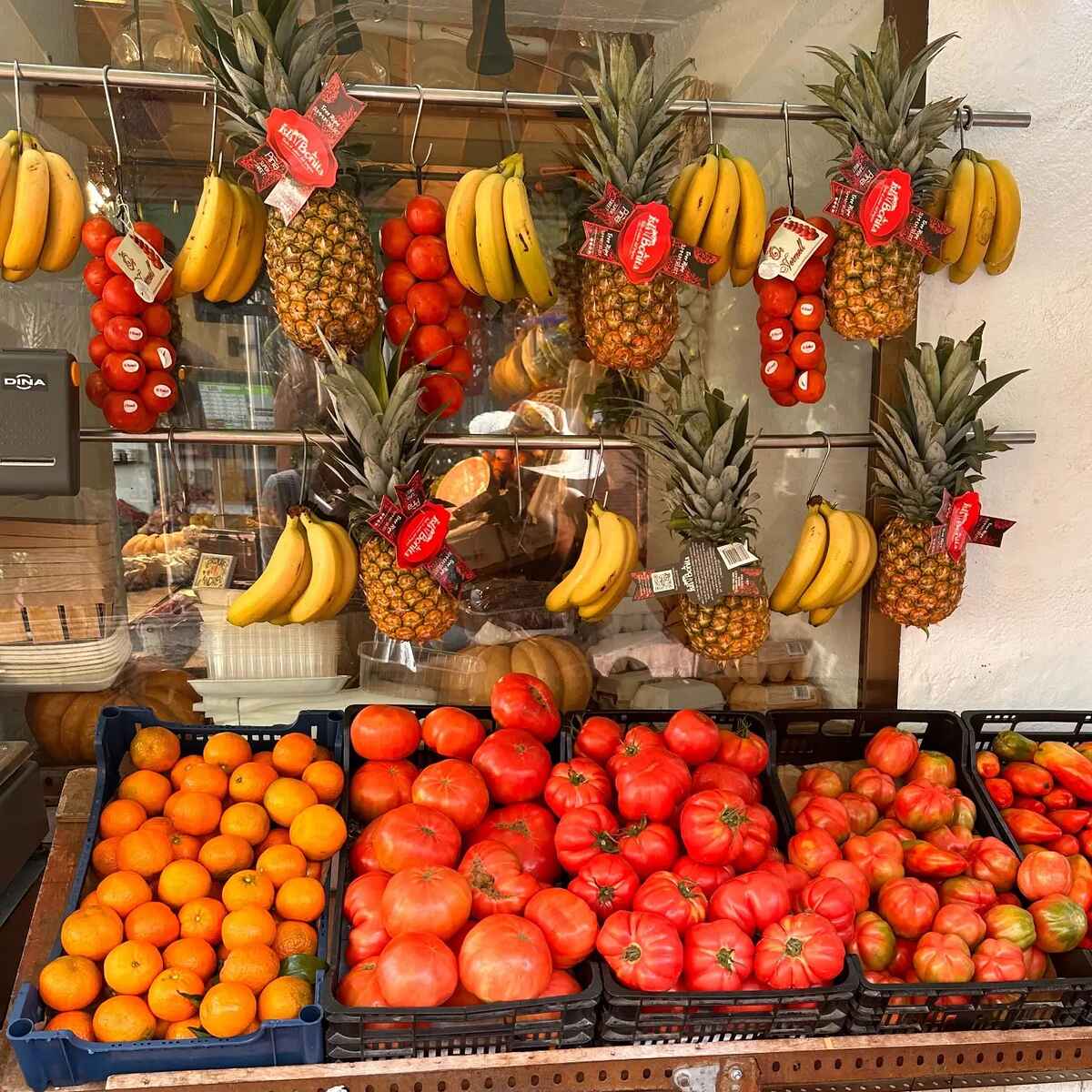 Vibrant produce display featuring hanging bunches of bananas and pineapples with red price tags, tomatoes, and oranges arranged in black crates below