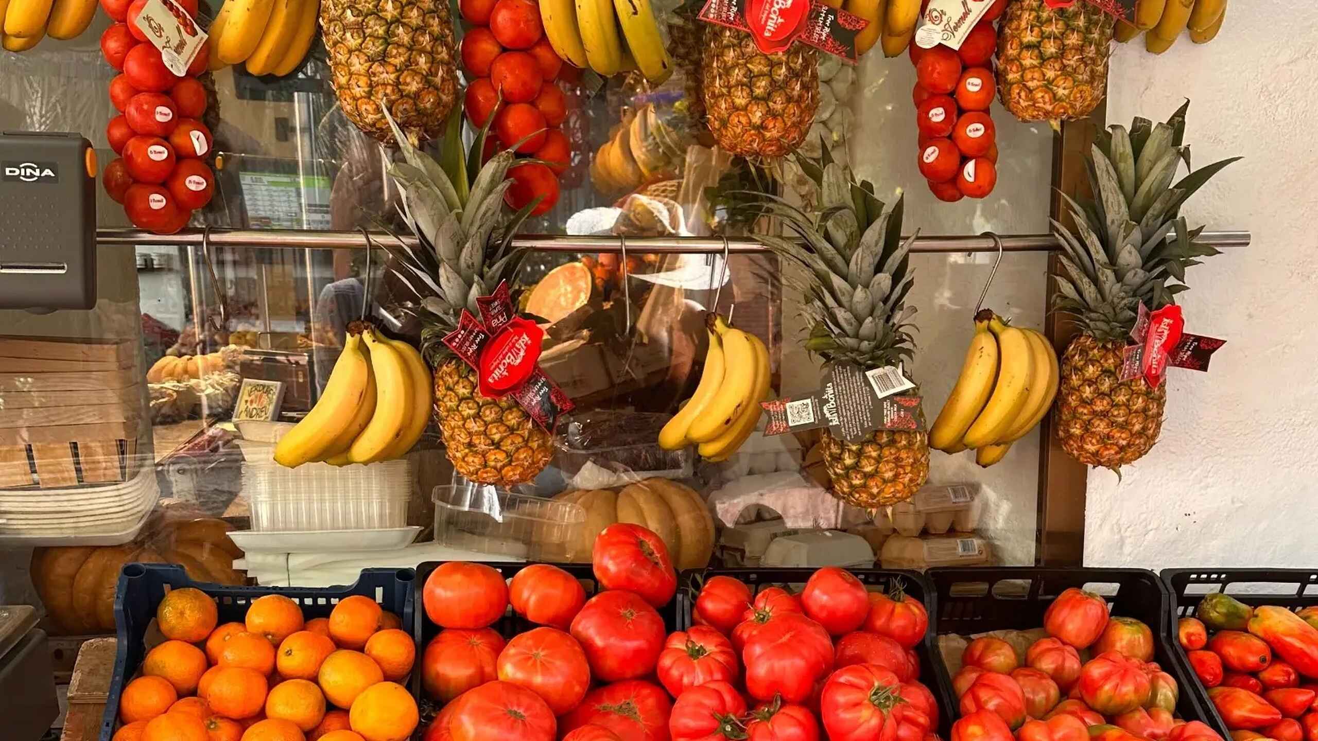 Vibrant produce display featuring hanging bunches of bananas and pineapples with red price tags, tomatoes, and oranges arranged in black crates below