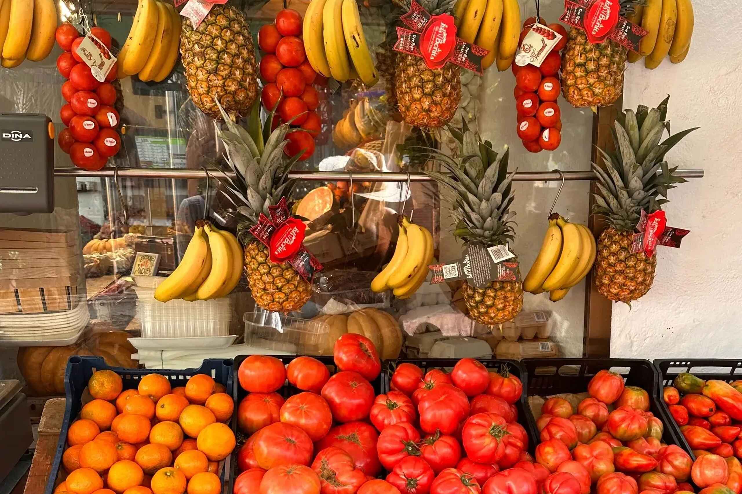 Vibrant produce display featuring hanging bunches of bananas and pineapples with red price tags, tomatoes, and oranges arranged in black crates below