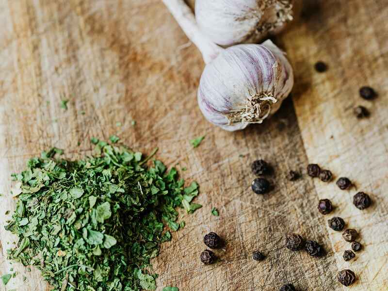 Garlic cloves and black peppercorns arranged on a wooden cutting board next to fresh herbs and ground spices for hemp pesto preparation