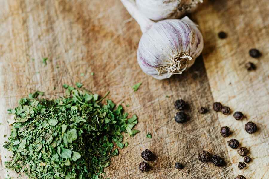 Garlic cloves and black peppercorns arranged on a wooden cutting board next to fresh herbs and ground spices for hemp pesto preparation