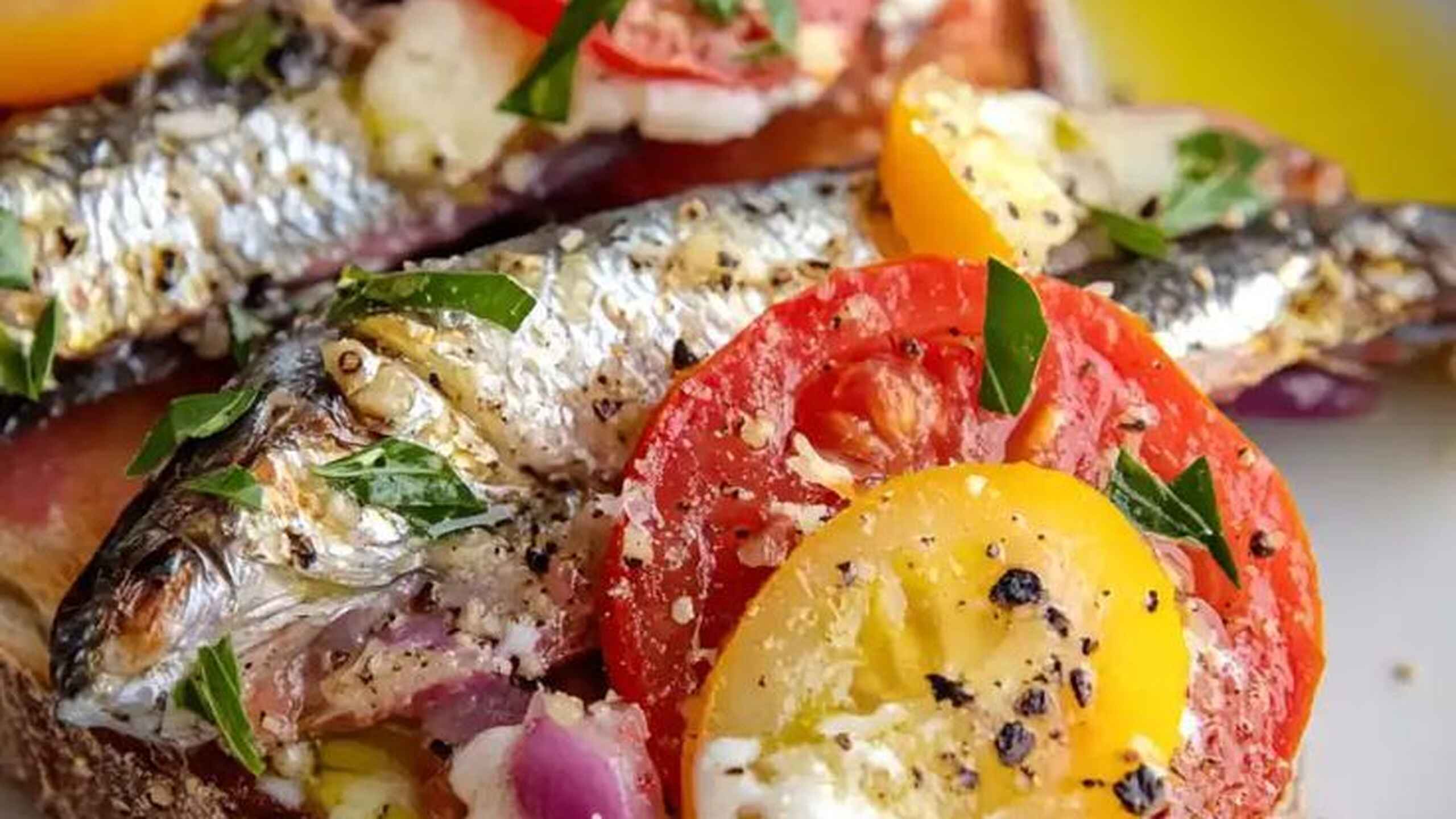 Sardines on toast topped with sliced tomatoes, yellow peppers, red onions, fresh chives, and creamy spread, drizzled with olive oil