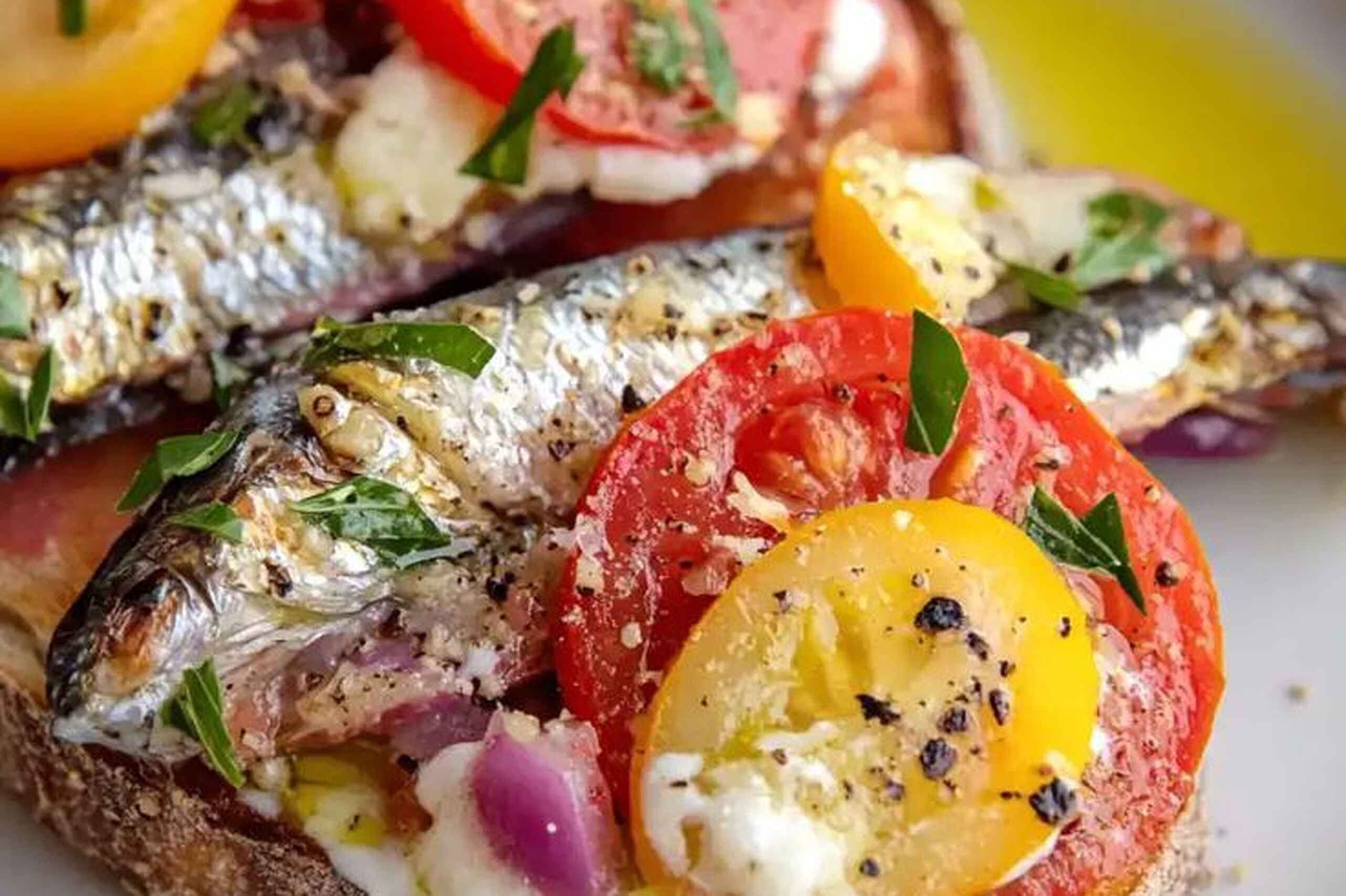 Sardines on toast topped with sliced tomatoes, yellow peppers, red onions, fresh chives, and creamy spread, drizzled with olive oil