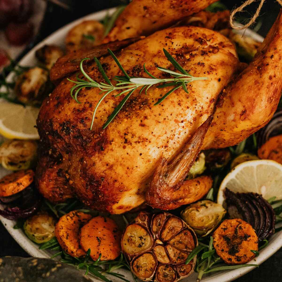 Whole roasted chicken with crispy golden skin garnished with fresh rosemary, surrounded by roasted garlic cloves, carrots, and lemon wedges on a white plate