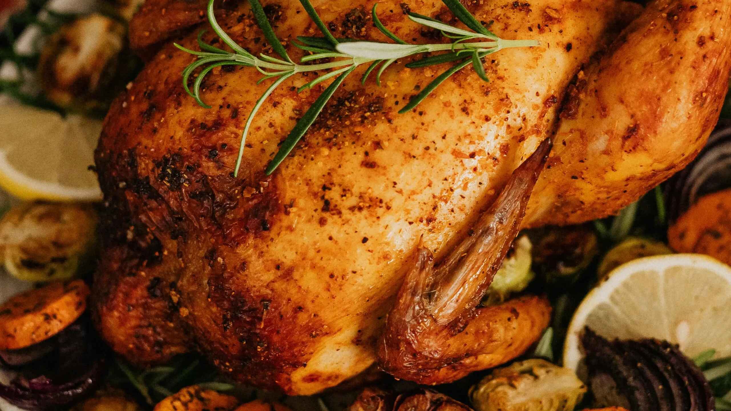 Whole roasted chicken with crispy golden skin garnished with fresh rosemary, surrounded by roasted garlic cloves, carrots, and lemon wedges on a white plate