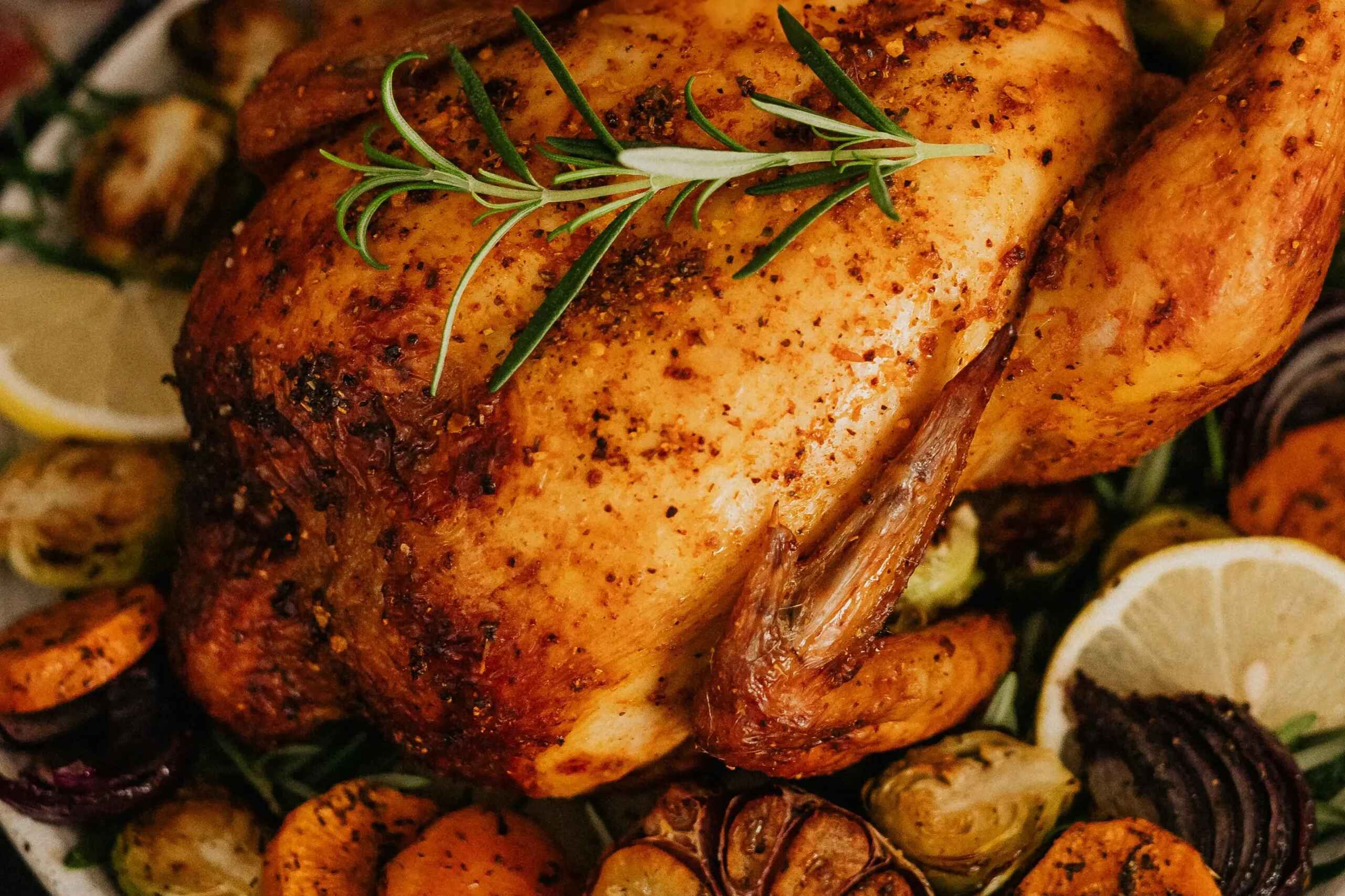 Whole roasted chicken with crispy golden skin garnished with fresh rosemary, surrounded by roasted garlic cloves, carrots, and lemon wedges on a white plate