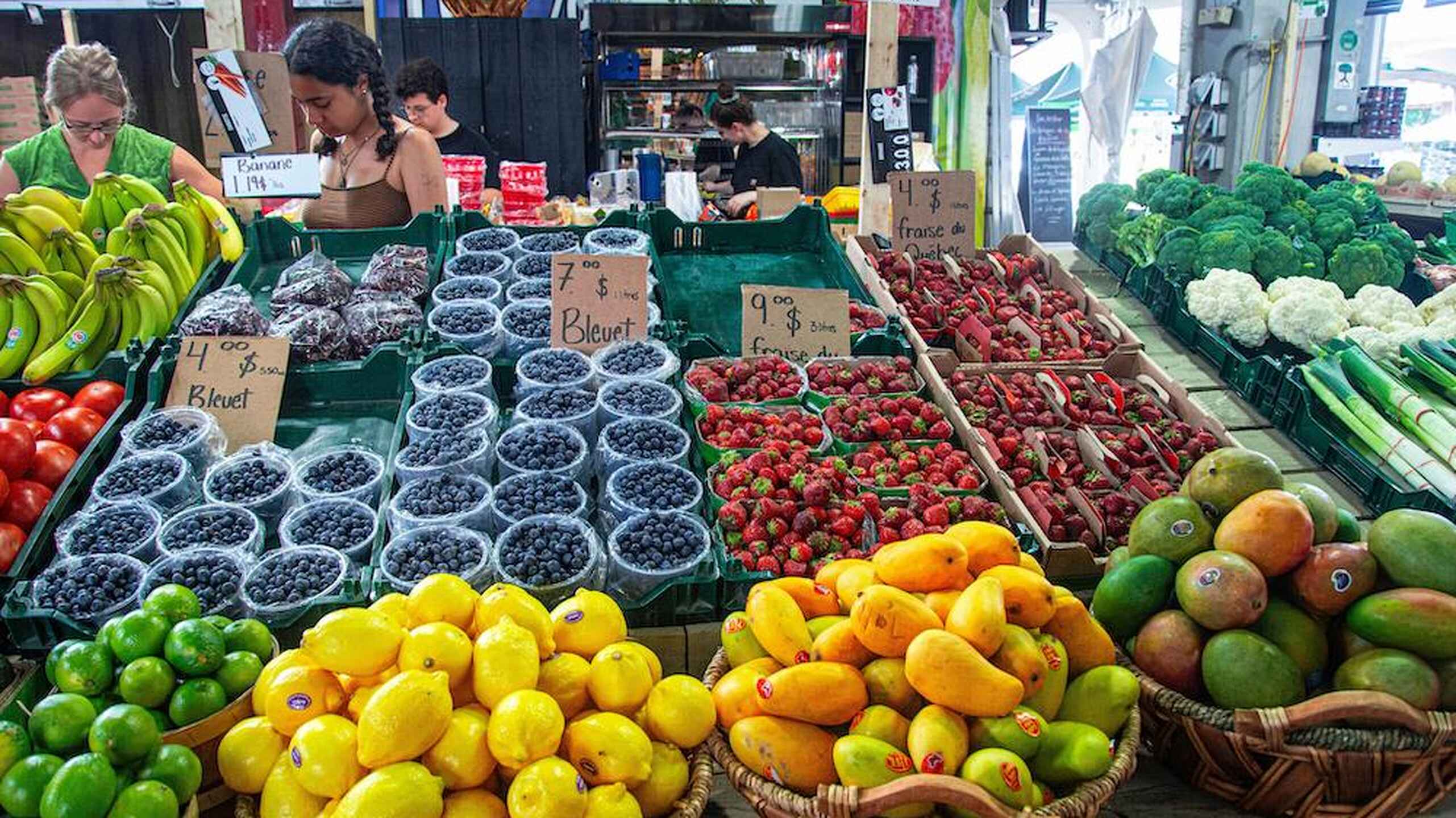 Farmer's market display overflowing with fresh produce including blueberries, strawberries, mangoes, citrus fruits, leafy greens, and cruciferous vegetables, with vendors at the counter
