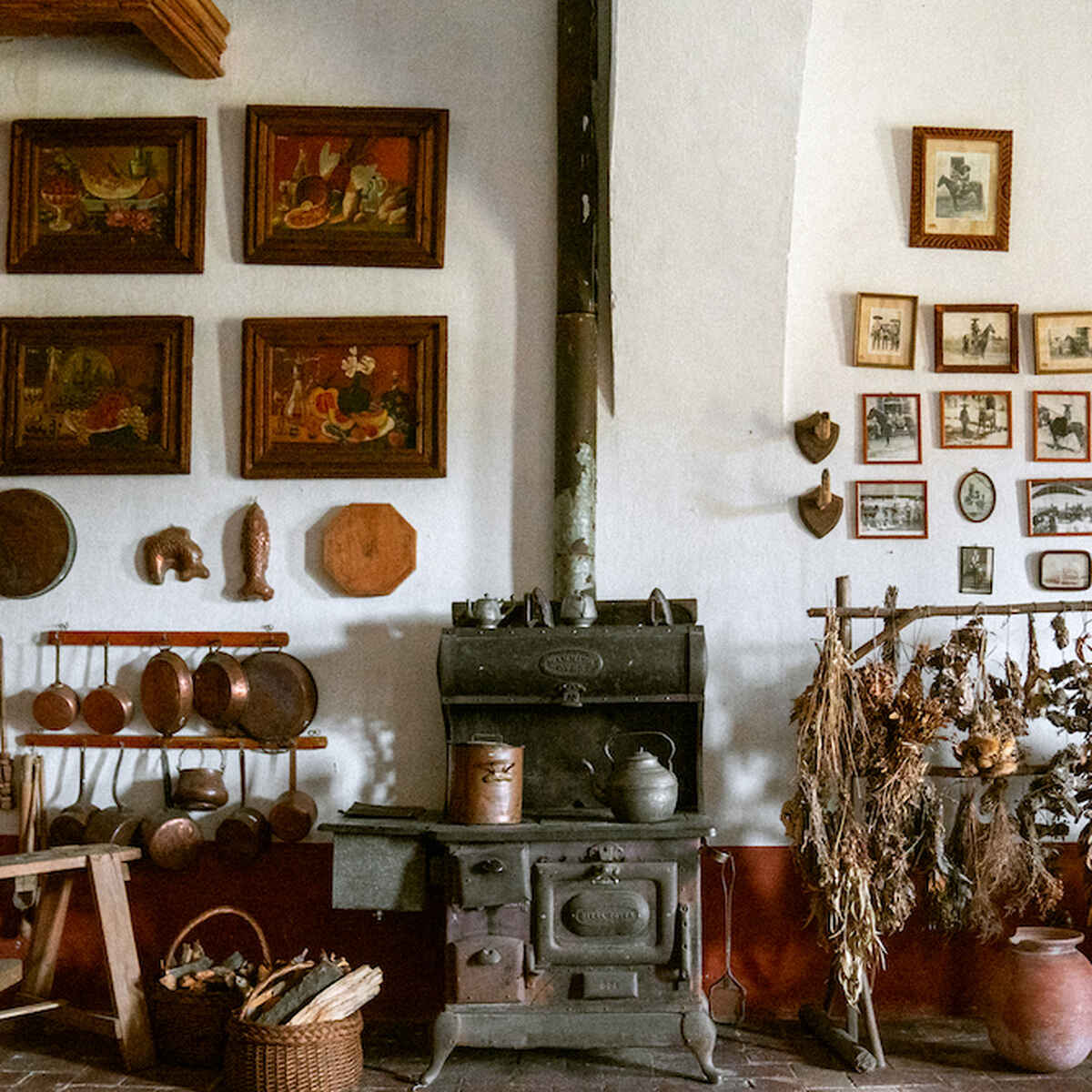 Historic farmhouse kitchen with cast iron cookware, a wood-burning stove, dried herbs and vegetables, and walls decorated with paintings and photographs