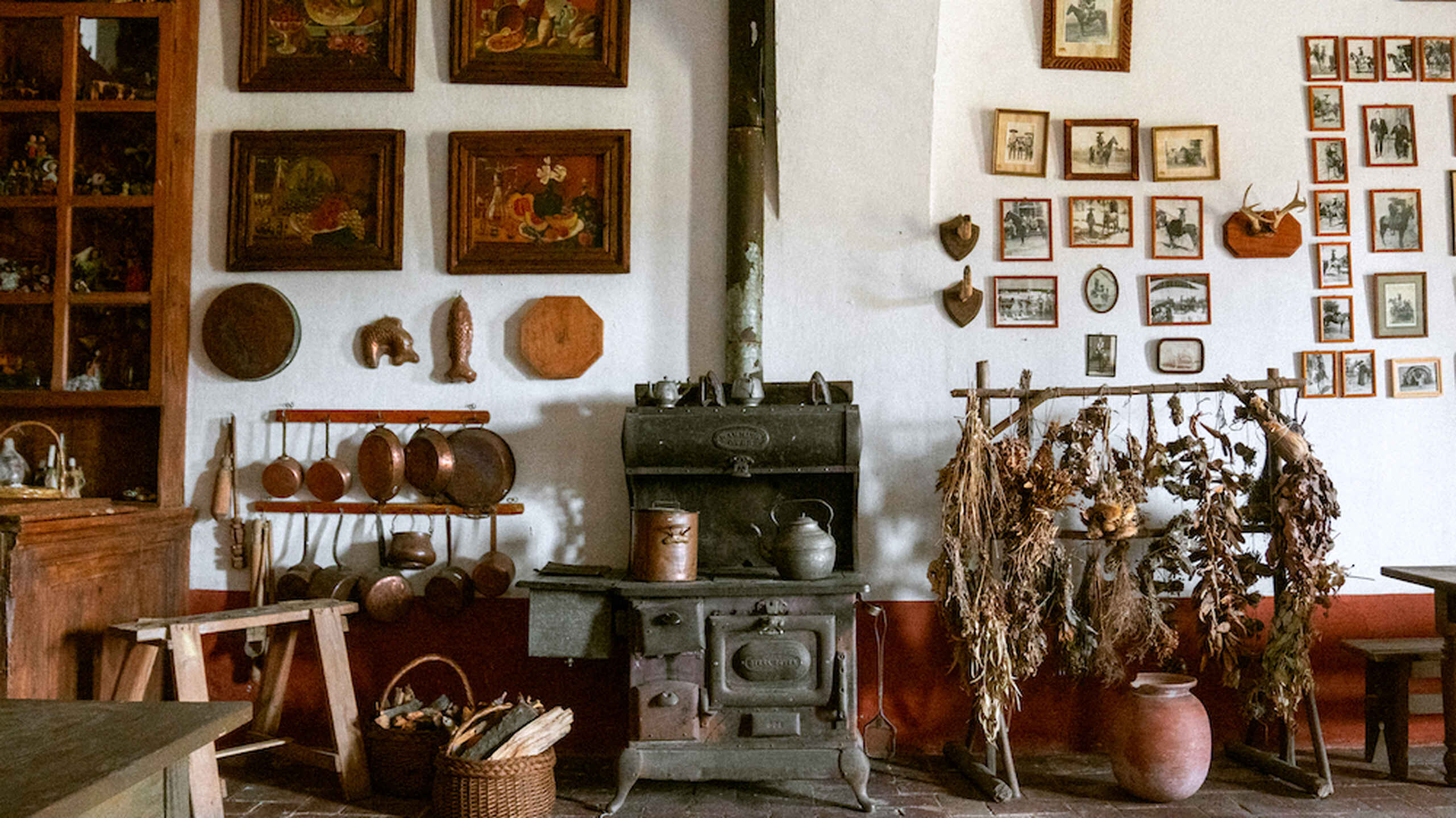 Historic farmhouse kitchen with cast iron cookware, a wood-burning stove, dried herbs and vegetables, and walls decorated with paintings and photographs