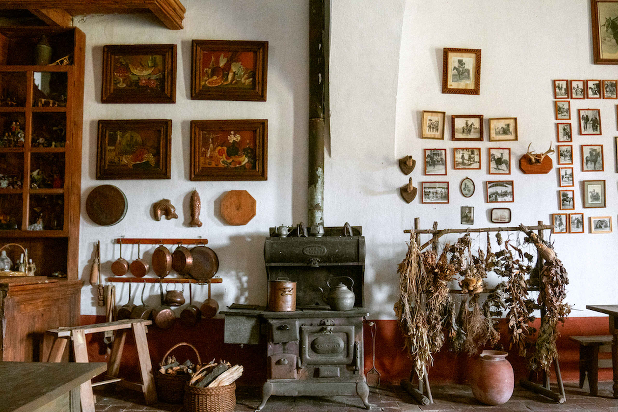 Historic farmhouse kitchen with cast iron cookware, a wood-burning stove, dried herbs and vegetables, and walls decorated with paintings and photographs