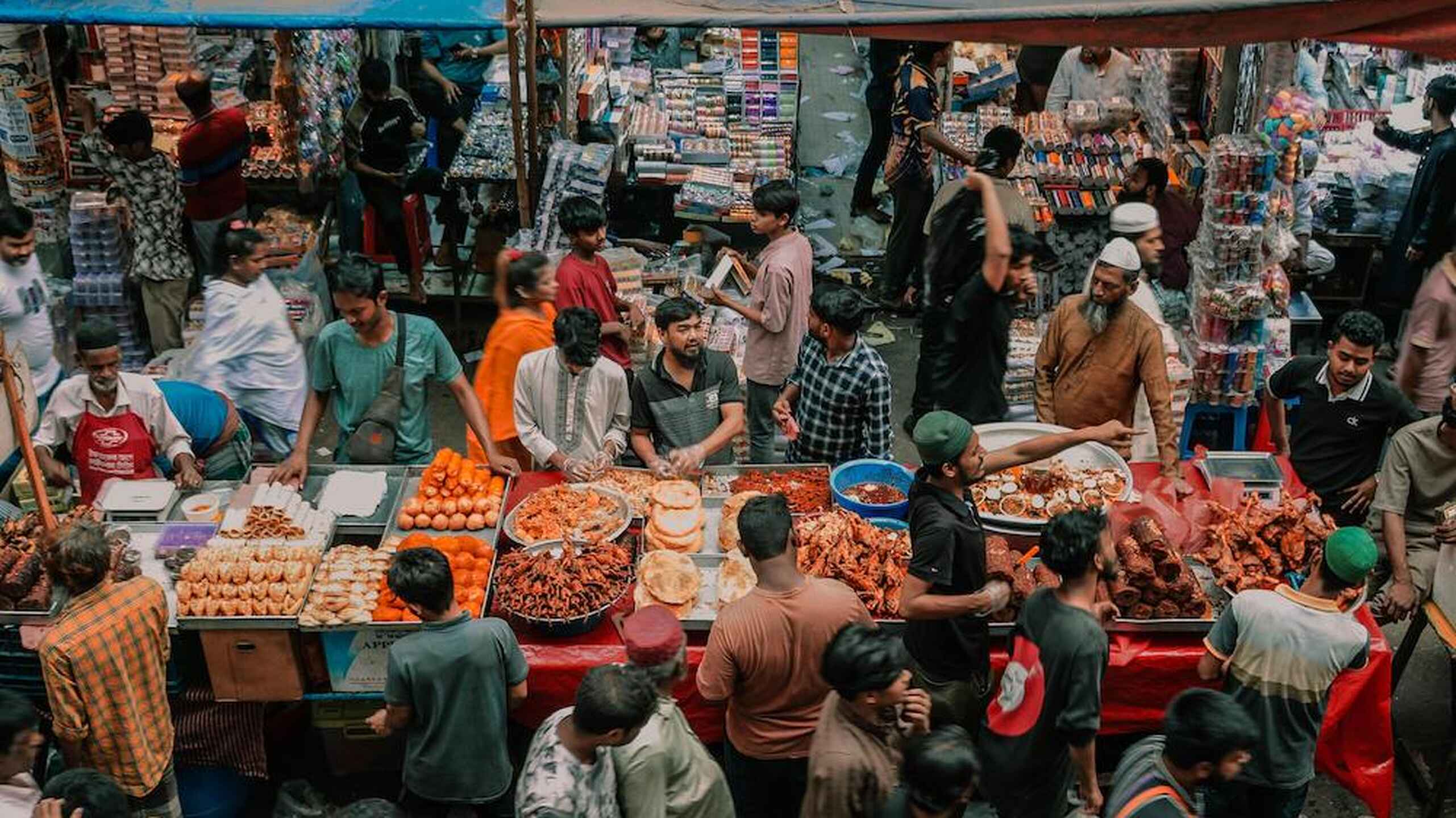 Crowded food market stall displaying fresh produce and prepared foods surrounded by shoppers and vendors