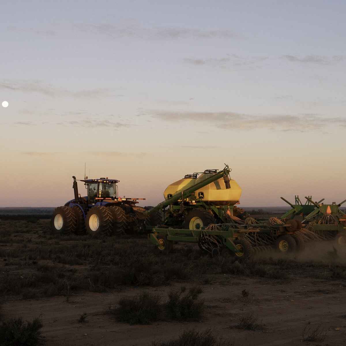 Large tractor pulling agricultural equipment across dusty farmland at dusk