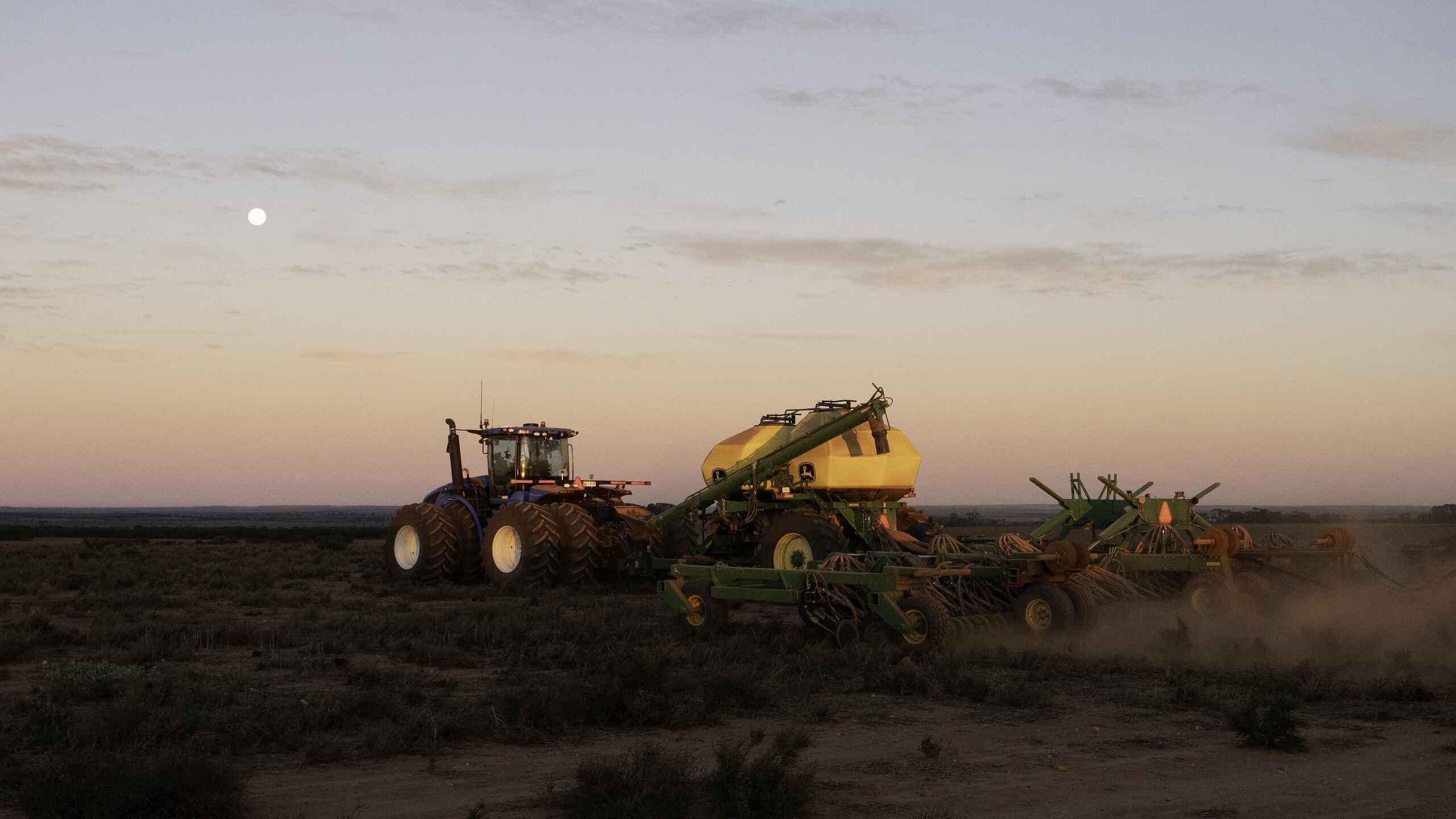Large tractor pulling agricultural equipment across dusty farmland at dusk