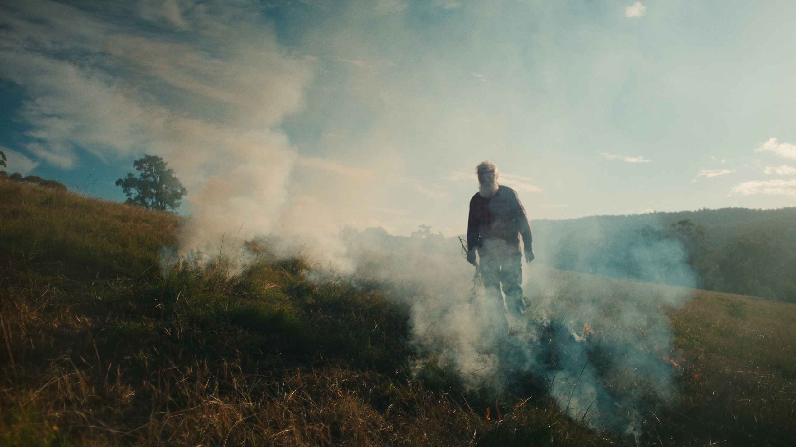 A farmer walks through smoke rising from a controlled burn in a dry grassland landscape