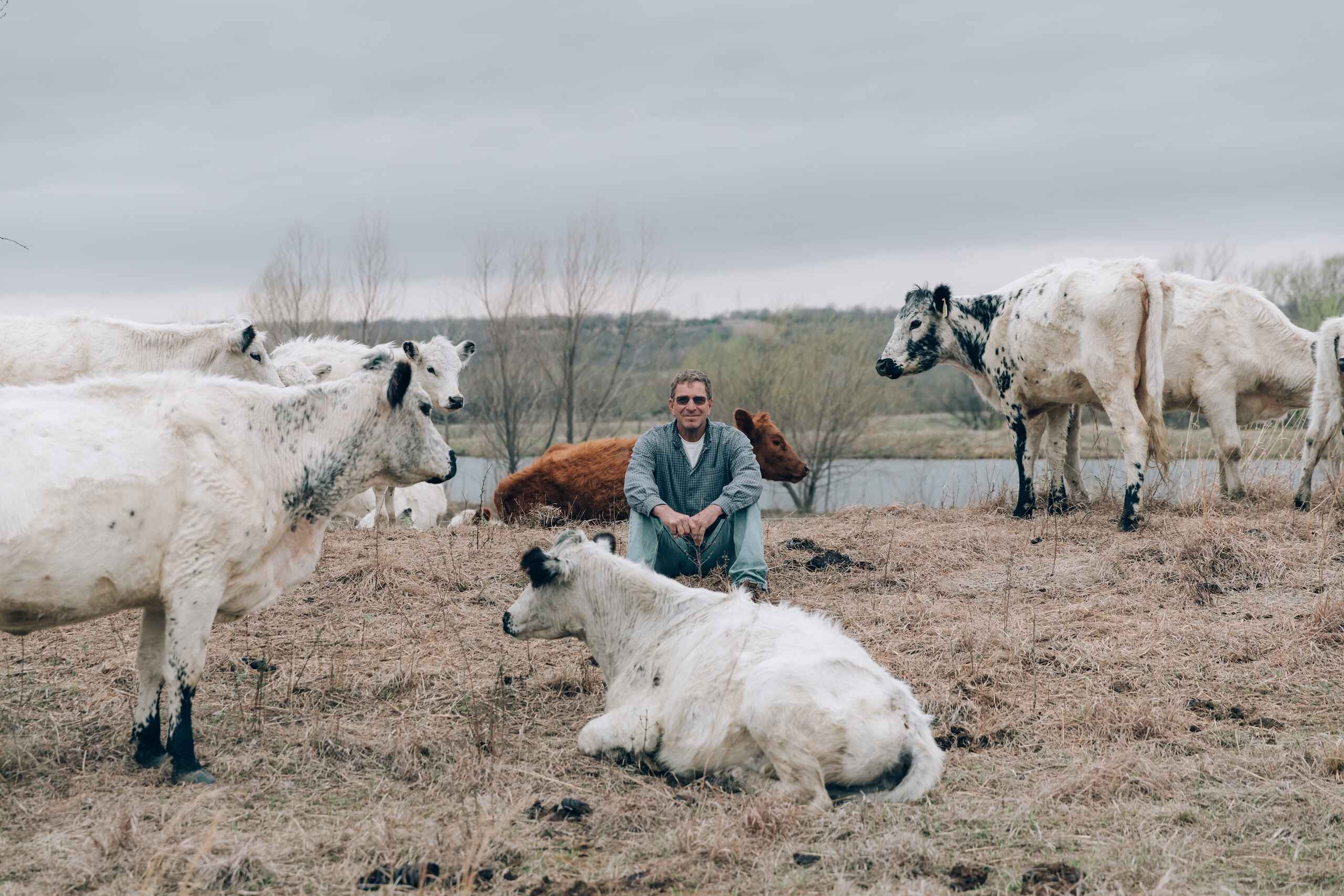 Farmer crouching among a herd of goats by a river in a regenerative farming setting