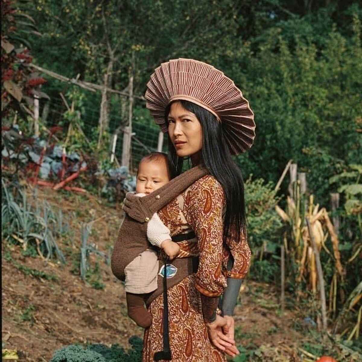 Woman holding a child in a vegetable garden with traditional clothing and a distinctive woven hat