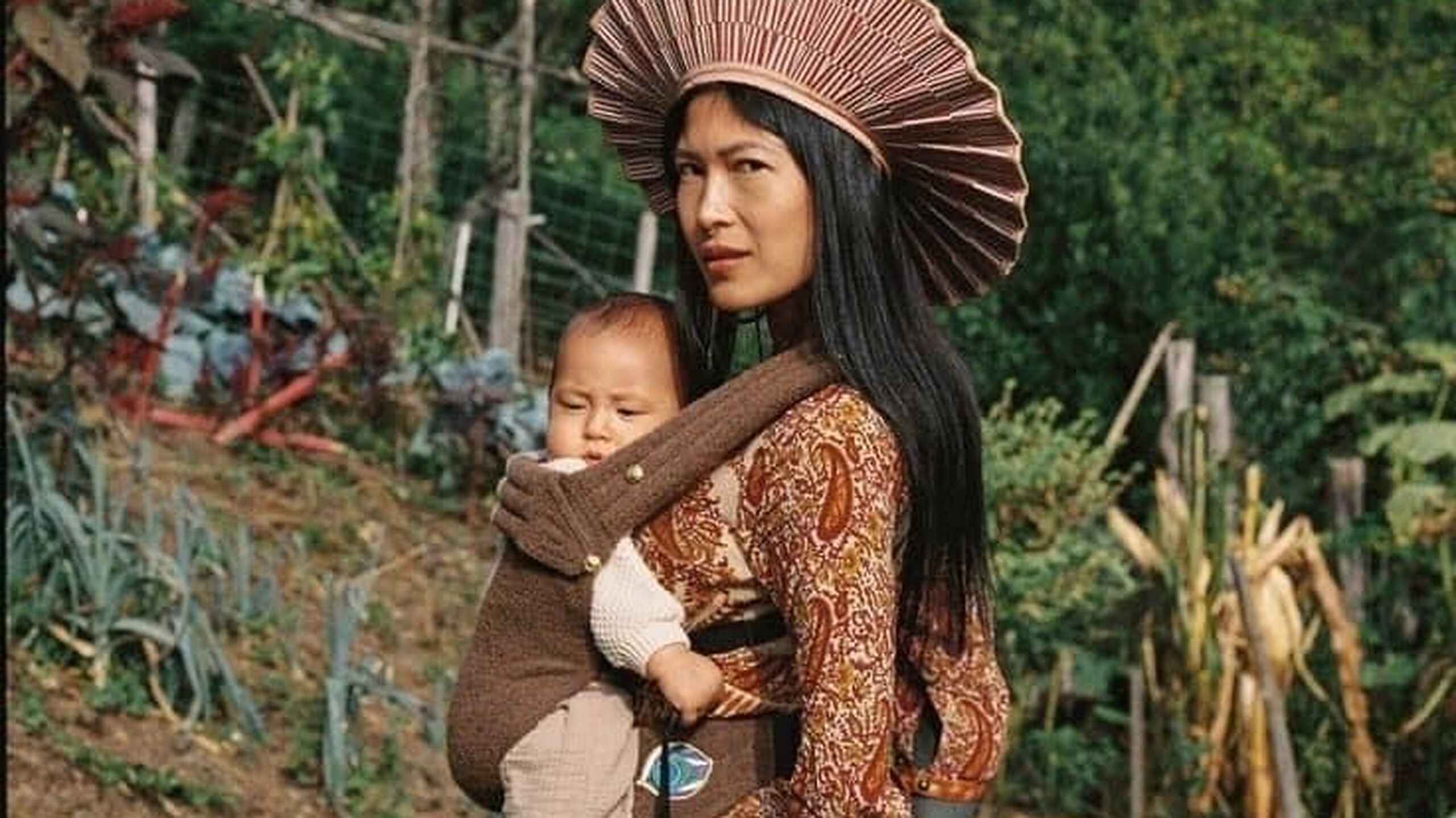 Woman holding a child in a vegetable garden with traditional clothing and a distinctive woven hat