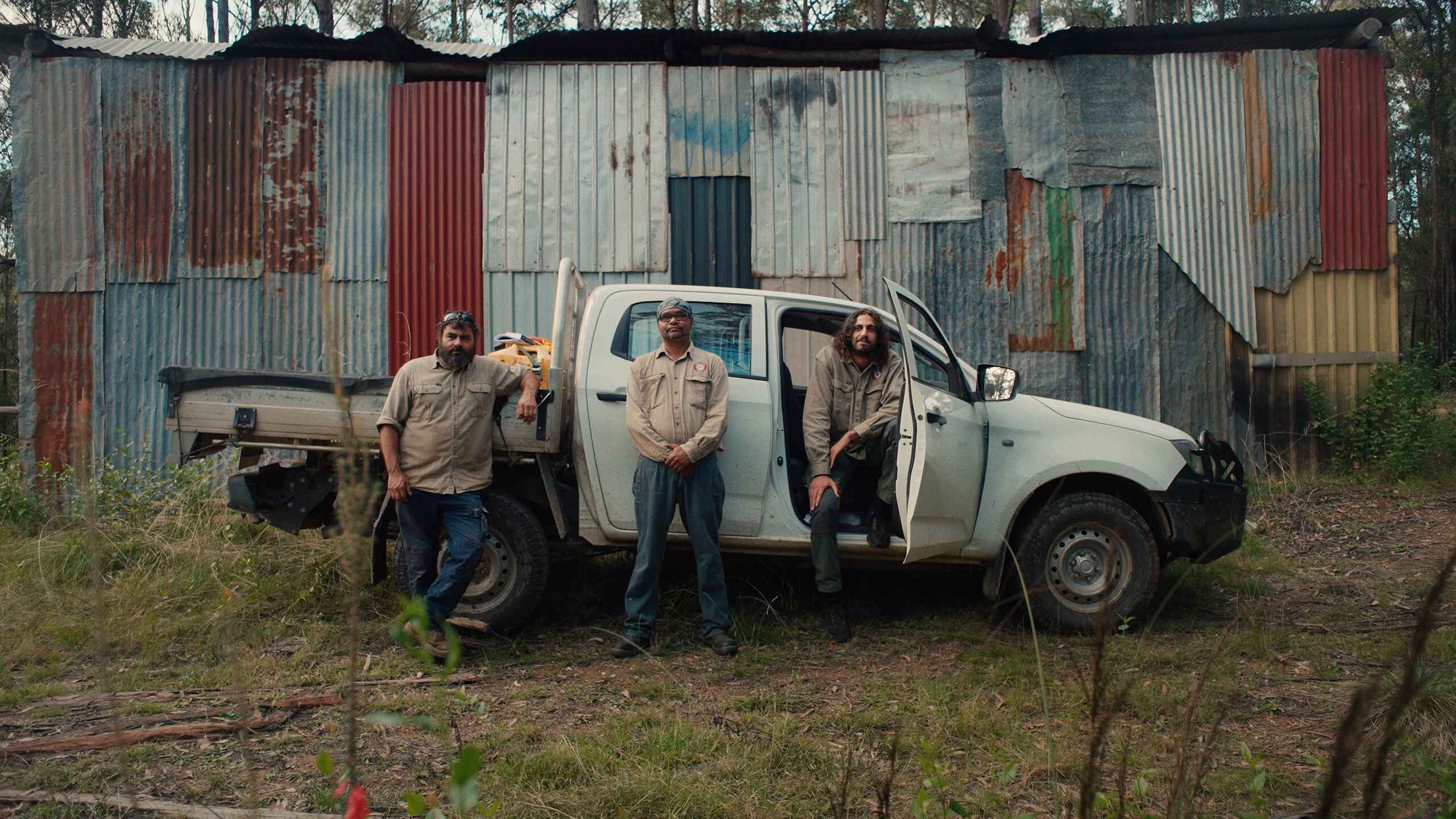 Three farmers stand in front of a white pickup truck and weathered corrugated metal building