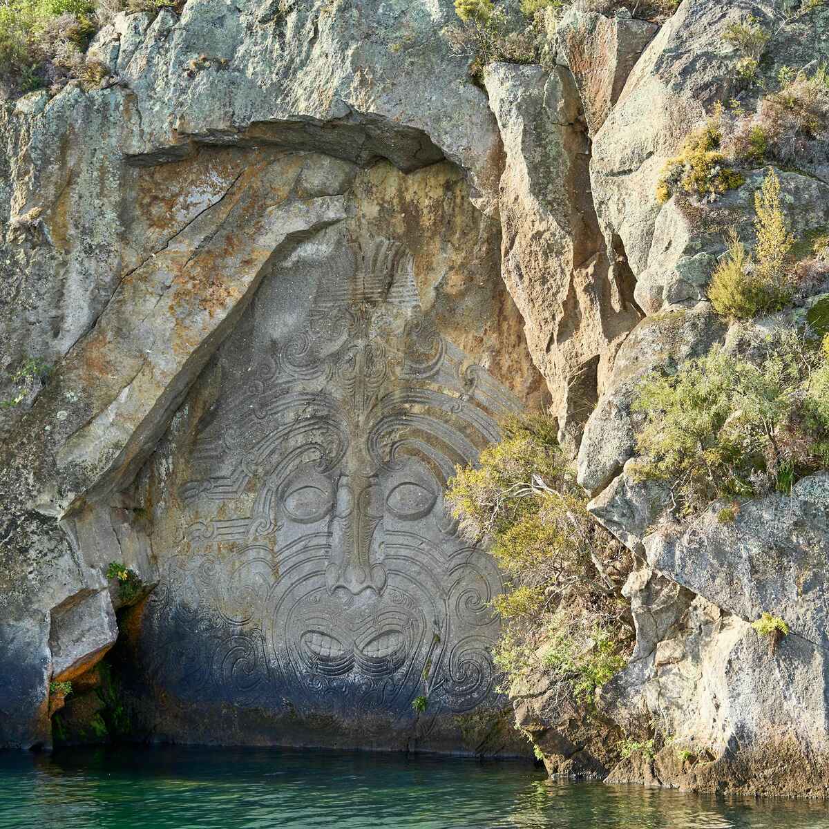 Maori carving of a face with intricate spiral patterns on a rock face above water