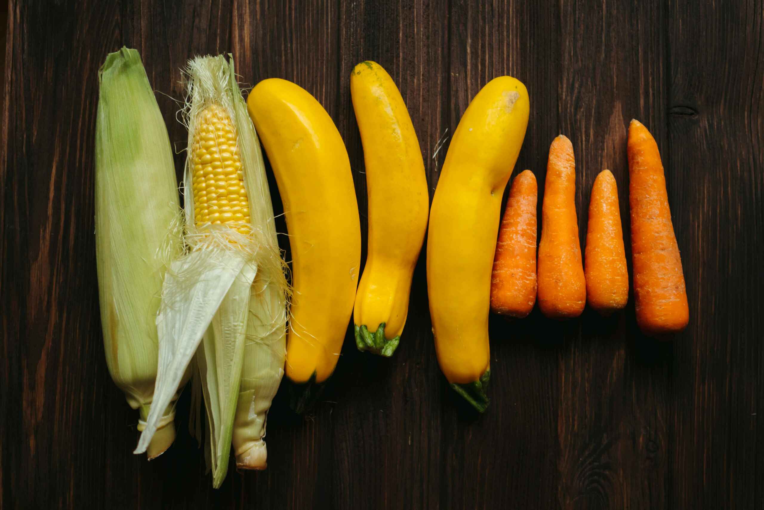 Yellow squash, corn, and carrots arranged in a row on a dark wooden surface