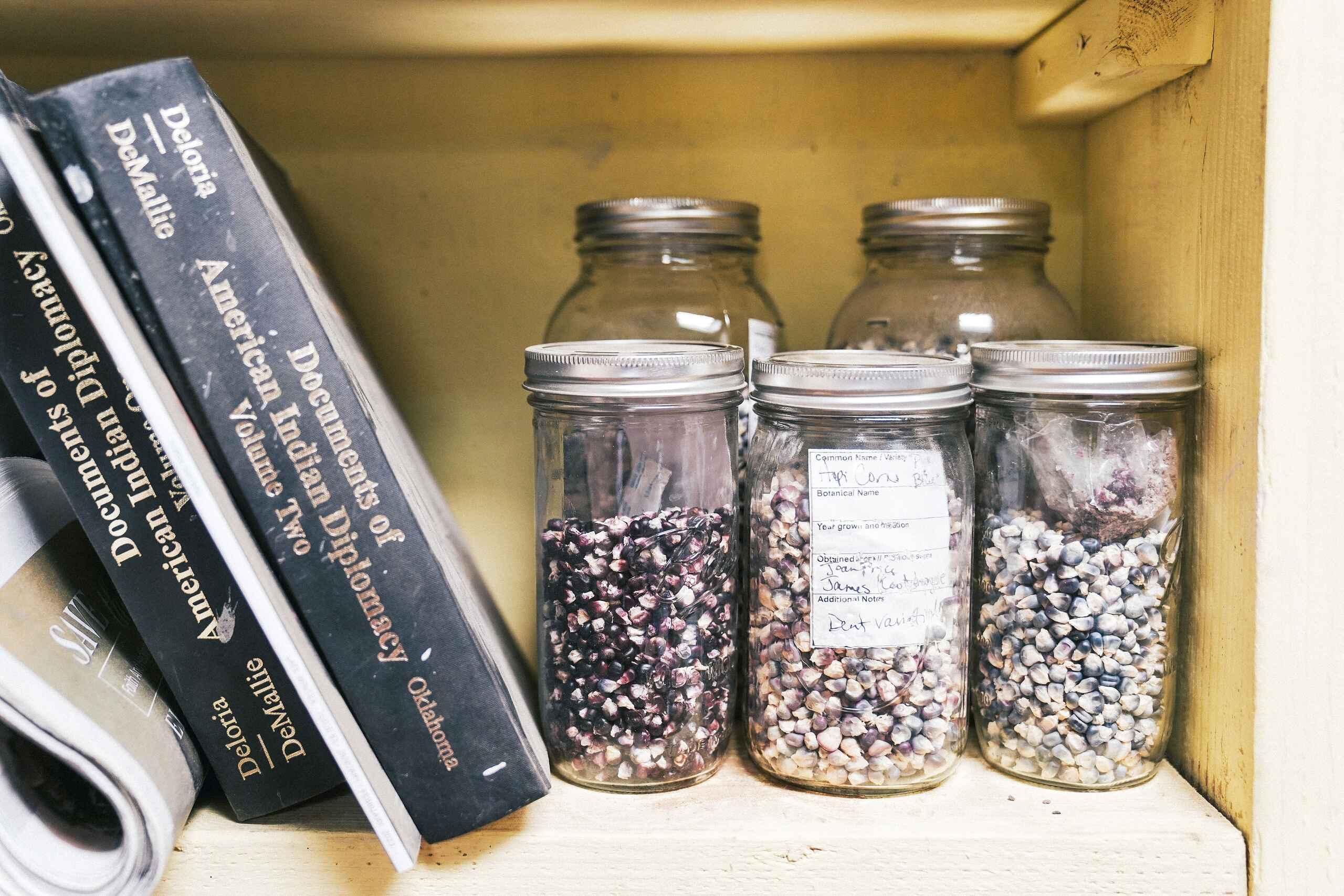 Wooden shelf with seed saving books and glass jars containing labeled Hopi crop varieties