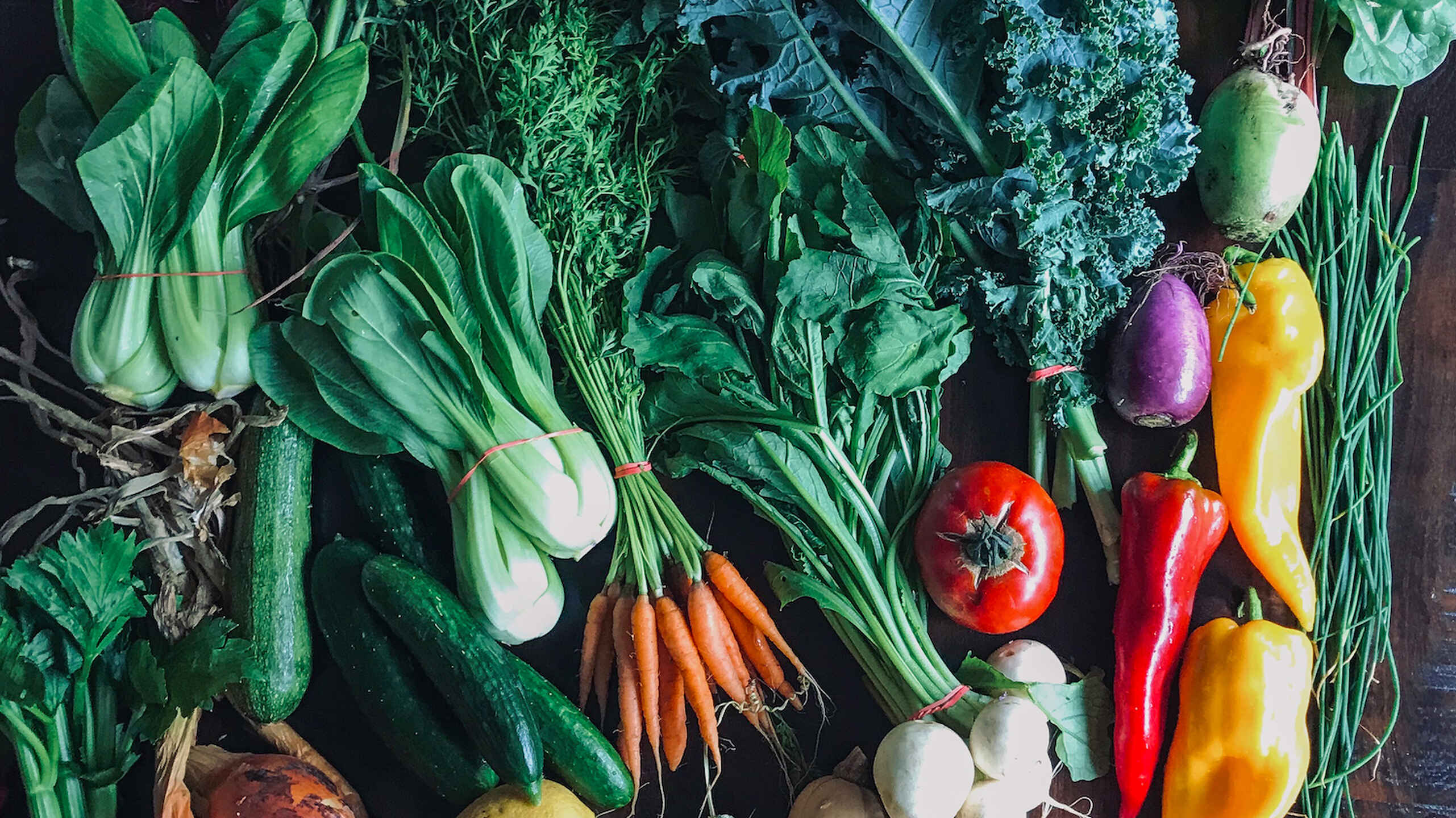 Fresh produce arranged on a dark surface including leafy greens, root vegetables, peppers, tomatoes, and squash