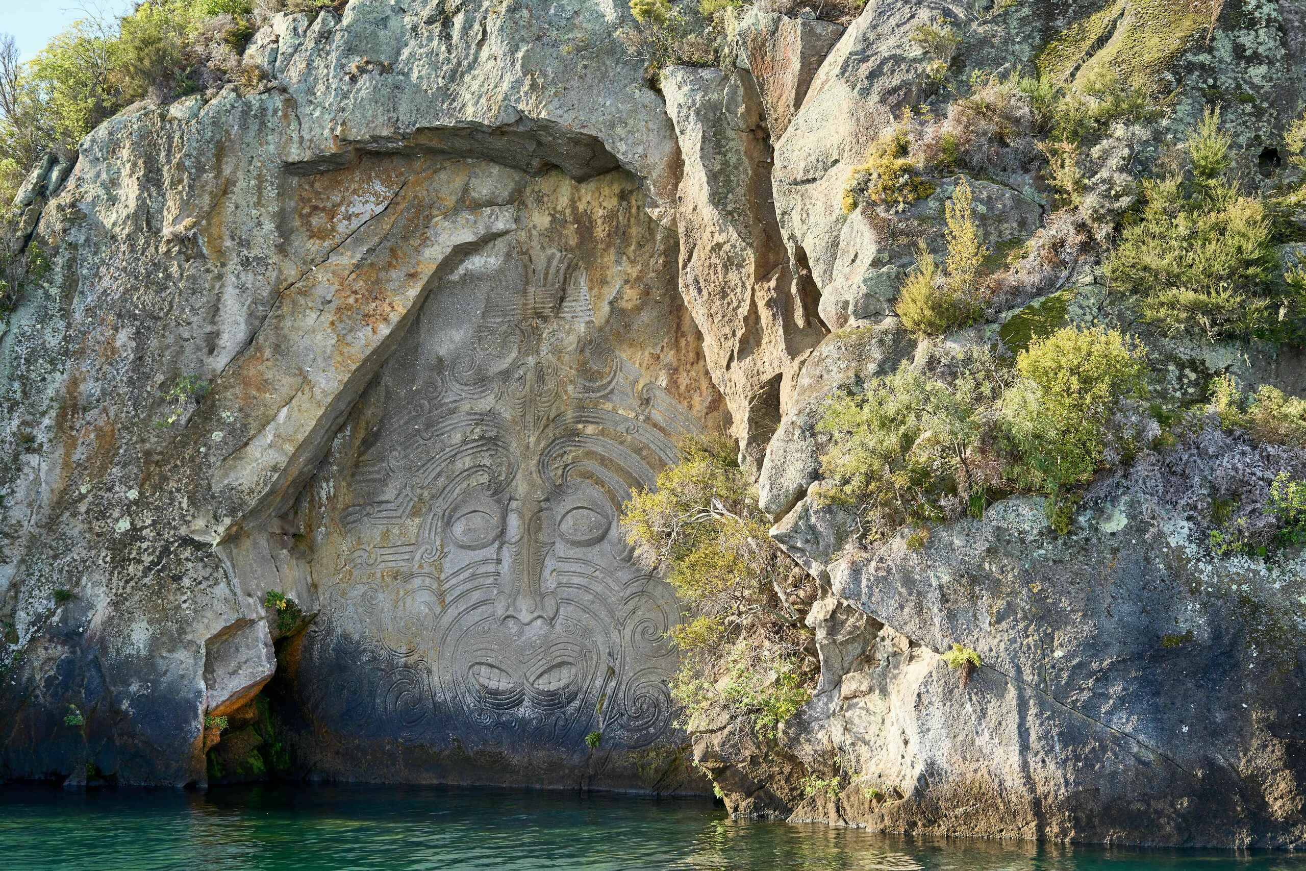 Maori carving of a face with traditional spiral patterns etched into a cliff face above turquoise water