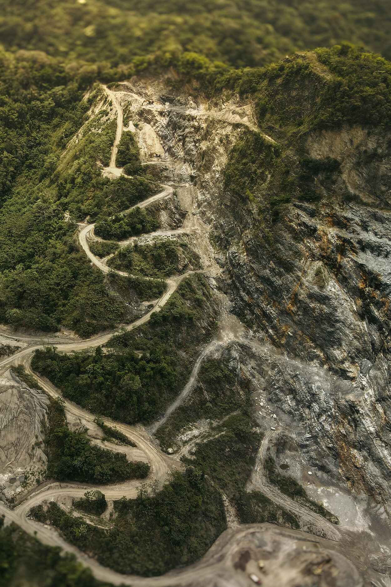 Aerial view of a winding mountain road carved through dense forest with exposed rocky terrain