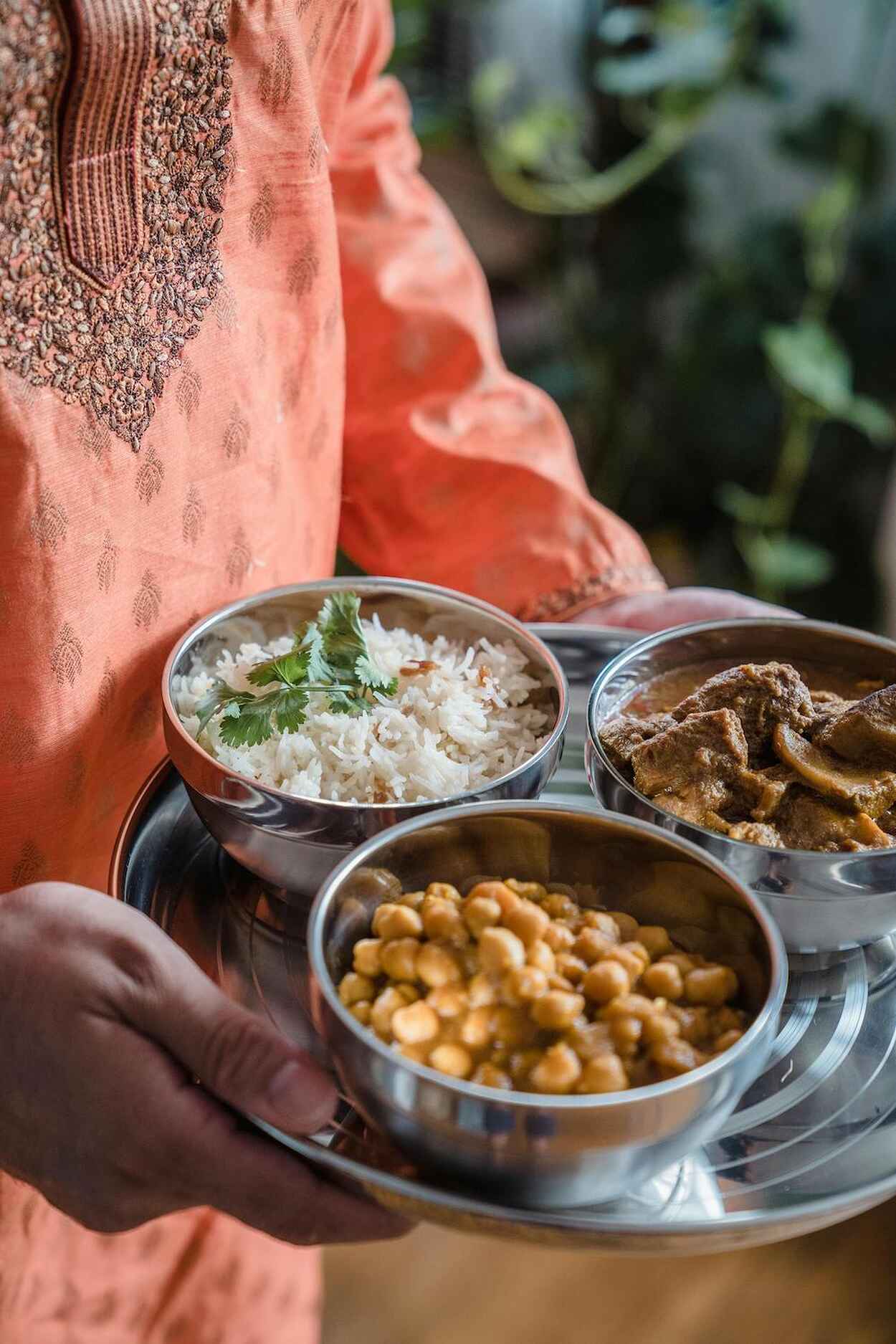Person in traditional South Asian dress holding a metal tray with rice, chickpeas, and meat curry