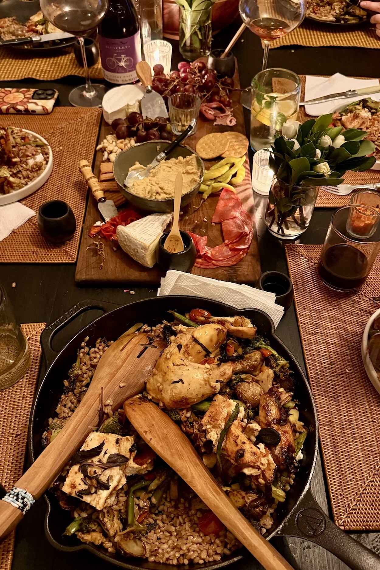 A dinner table spread with roasted chicken and grains in a cast iron skillet, surrounded by cheese, cured meats, fresh flowers, wine, and various prepared dishes