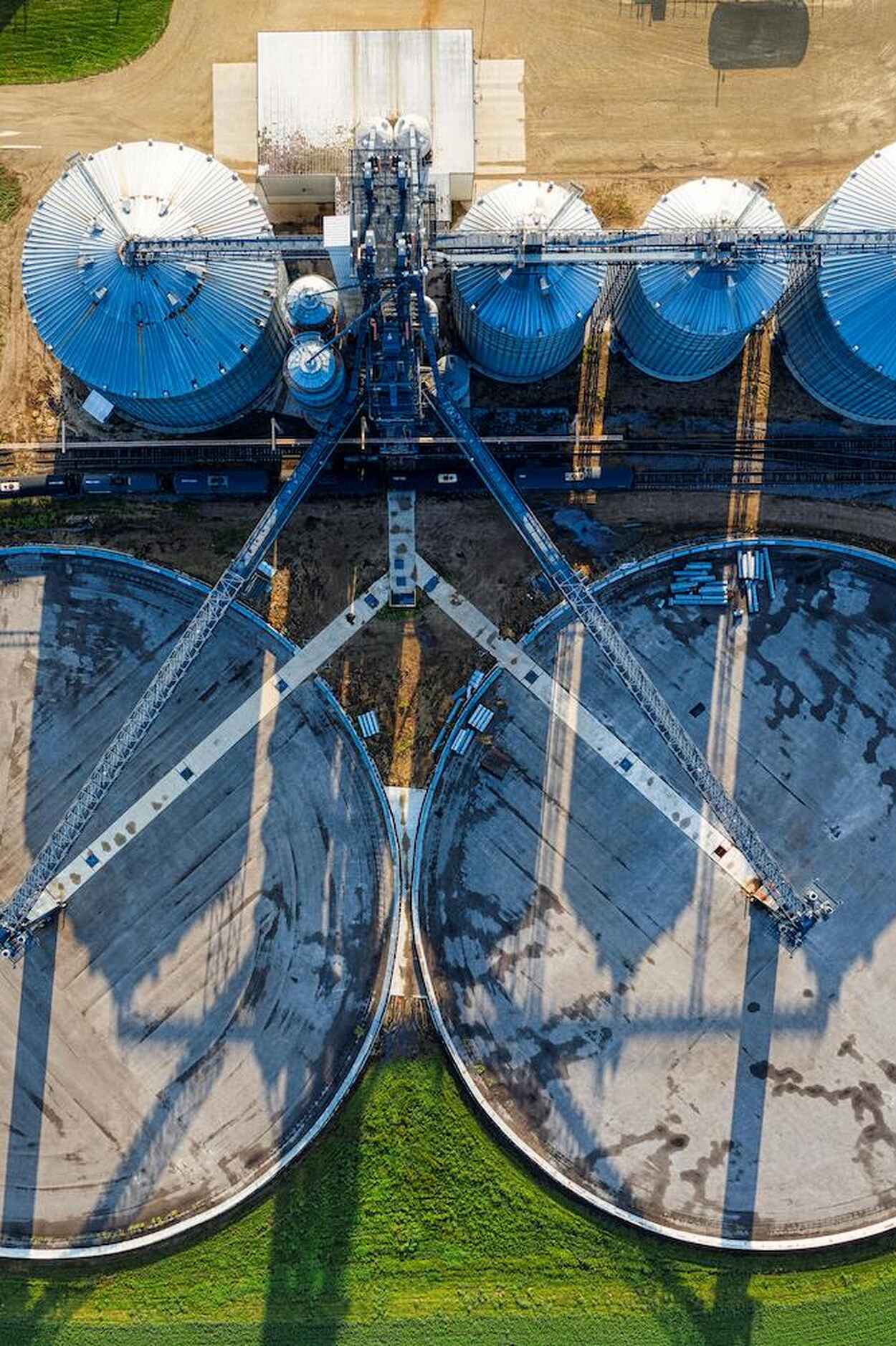 Aerial view of a large-scale grain storage facility with multiple blue and white cylindrical silos connected by conveyors and surrounded by two massive circular grain bins