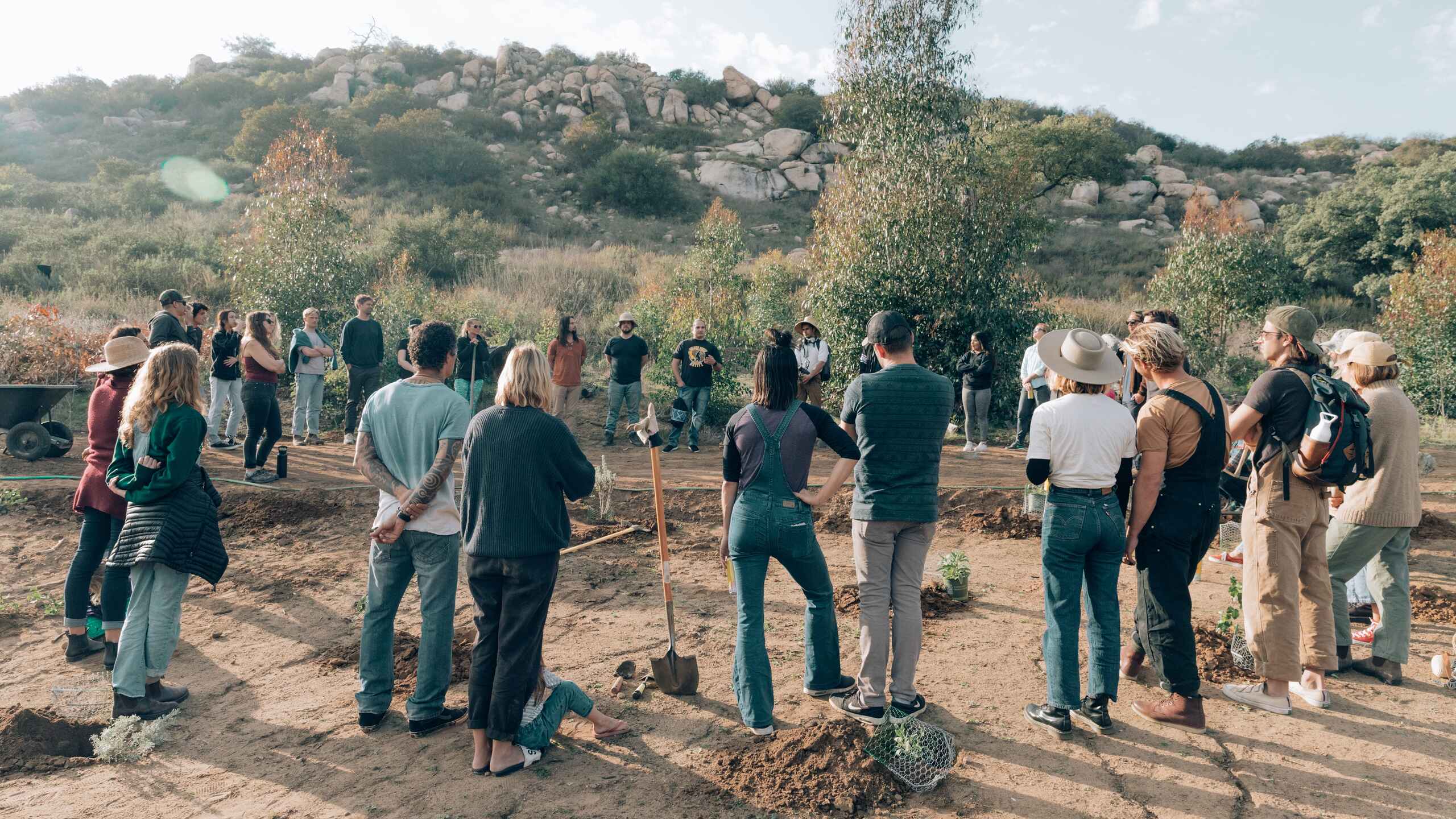 Community members gather for a tree-planting event in a natural landscape with rolling hills and rock formations