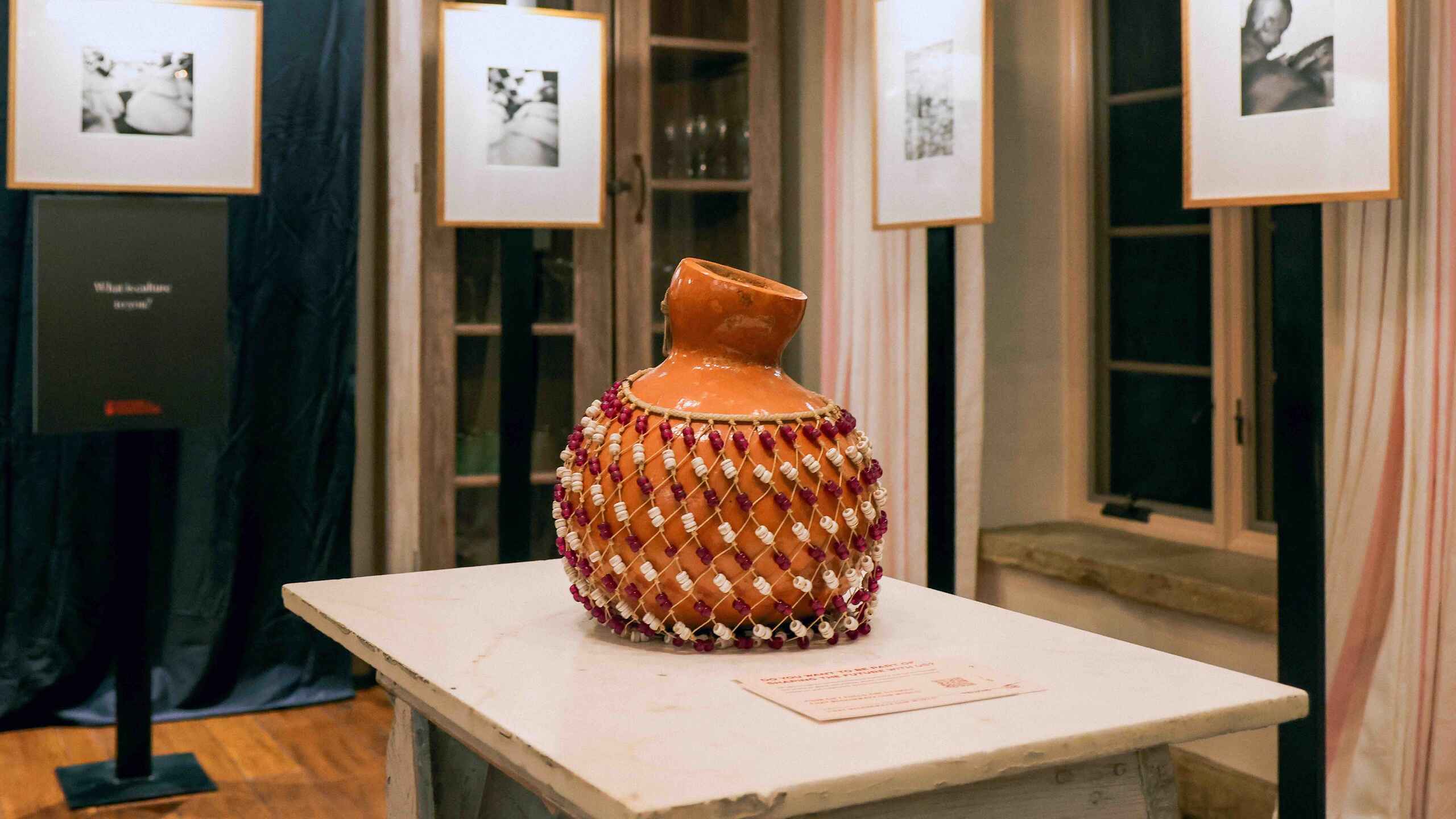 Terracotta vessel with woven red and pink fiber wrapping displayed on a pedestal in an art exhibition gallery with framed black and white photographs on the walls