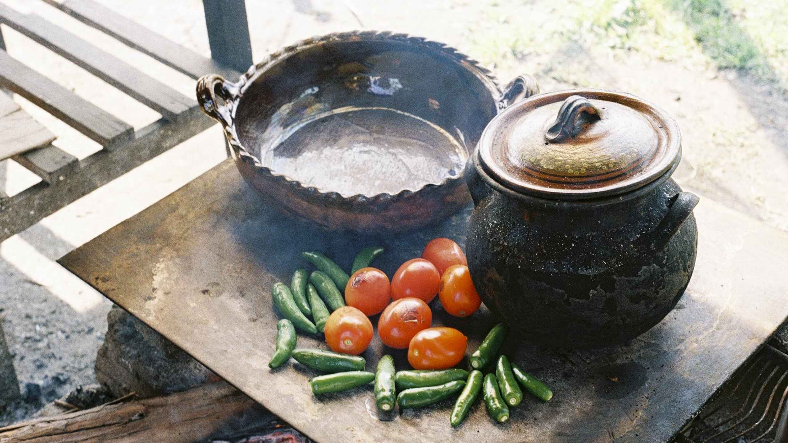 Cast iron cookware and fresh tomatoes and okra arranged on a stone surface over a fire, representing traditional cooking methods