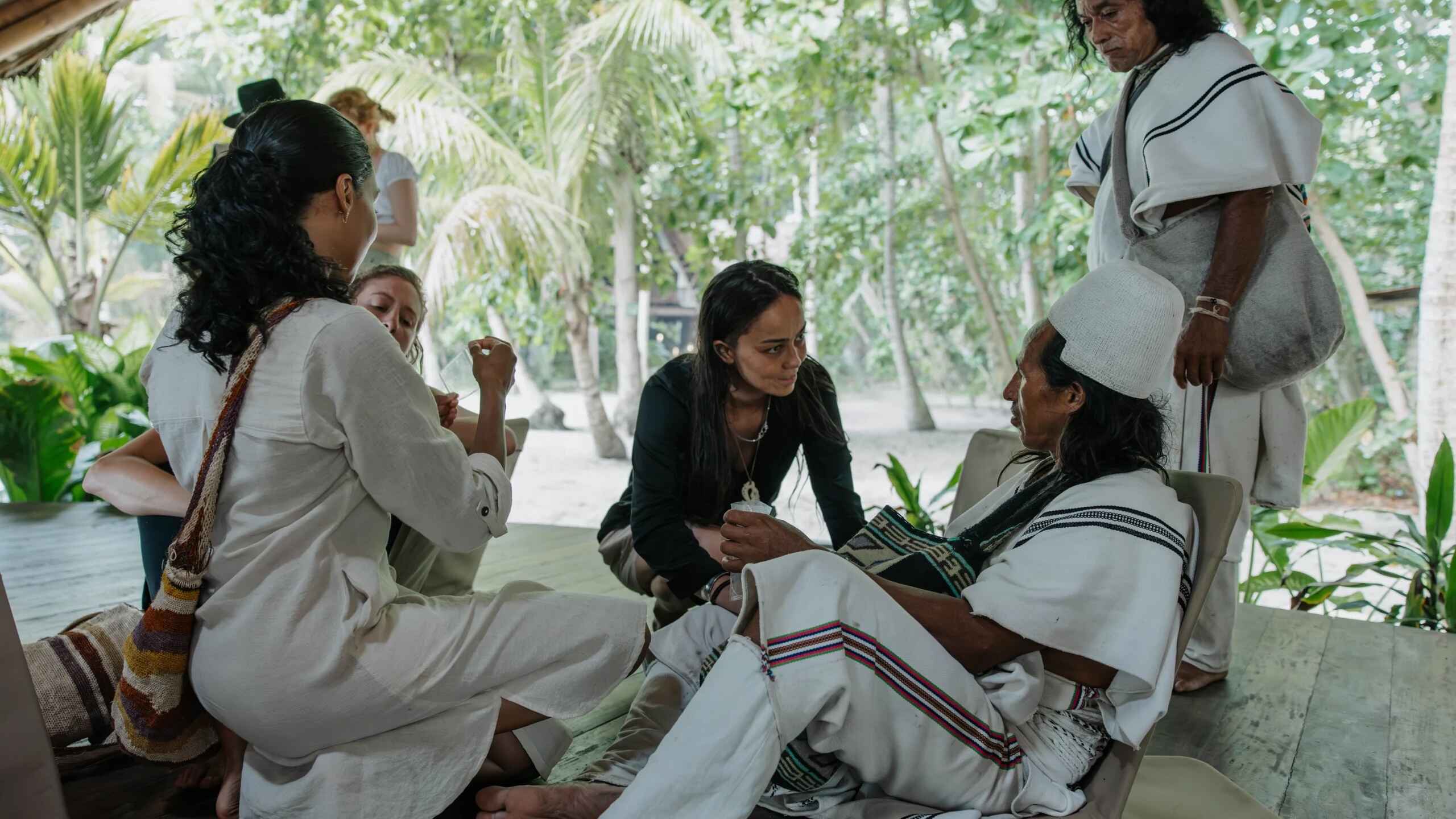 Indigenous woman in black shirt engages in discussion with community elders wearing traditional white clothing in a palm-thatched gathering space