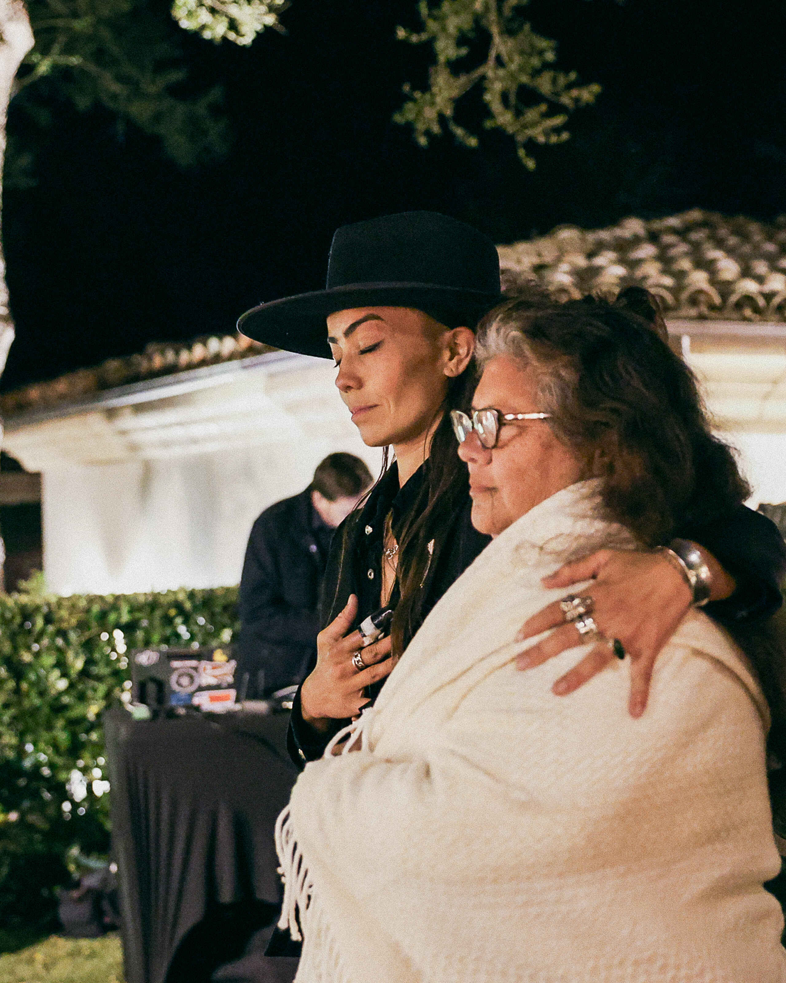 Two women embrace at a nighttime outdoor gathering, one wearing a black hat and the other in glasses and a cream-colored shawl
