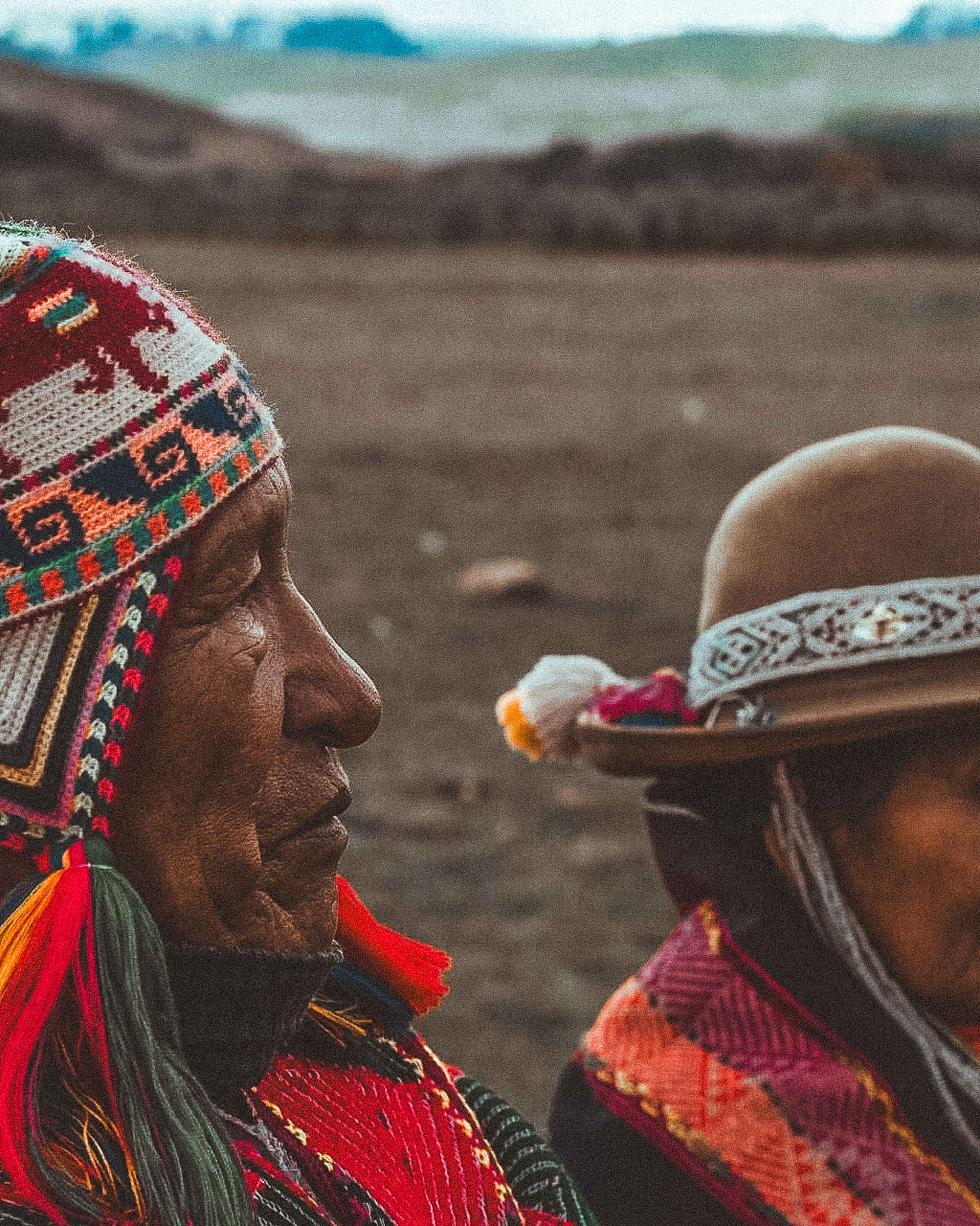 Two indigenous Andean elders wearing traditional colorful woven textiles and headwear stand together against a mountainous landscape