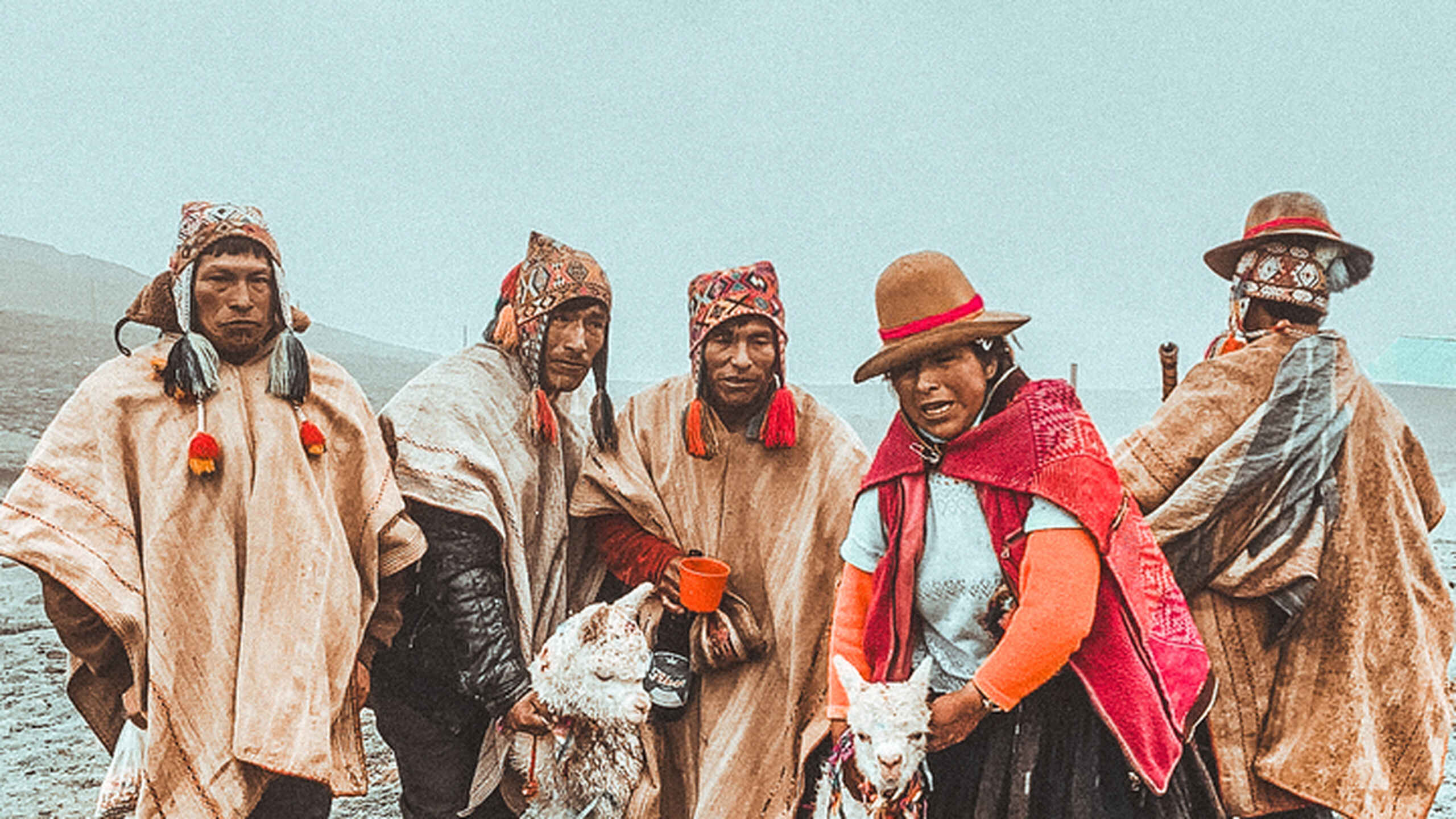 Five Peruvian elders in traditional clothing and alpacas stand together by a mountain lake
