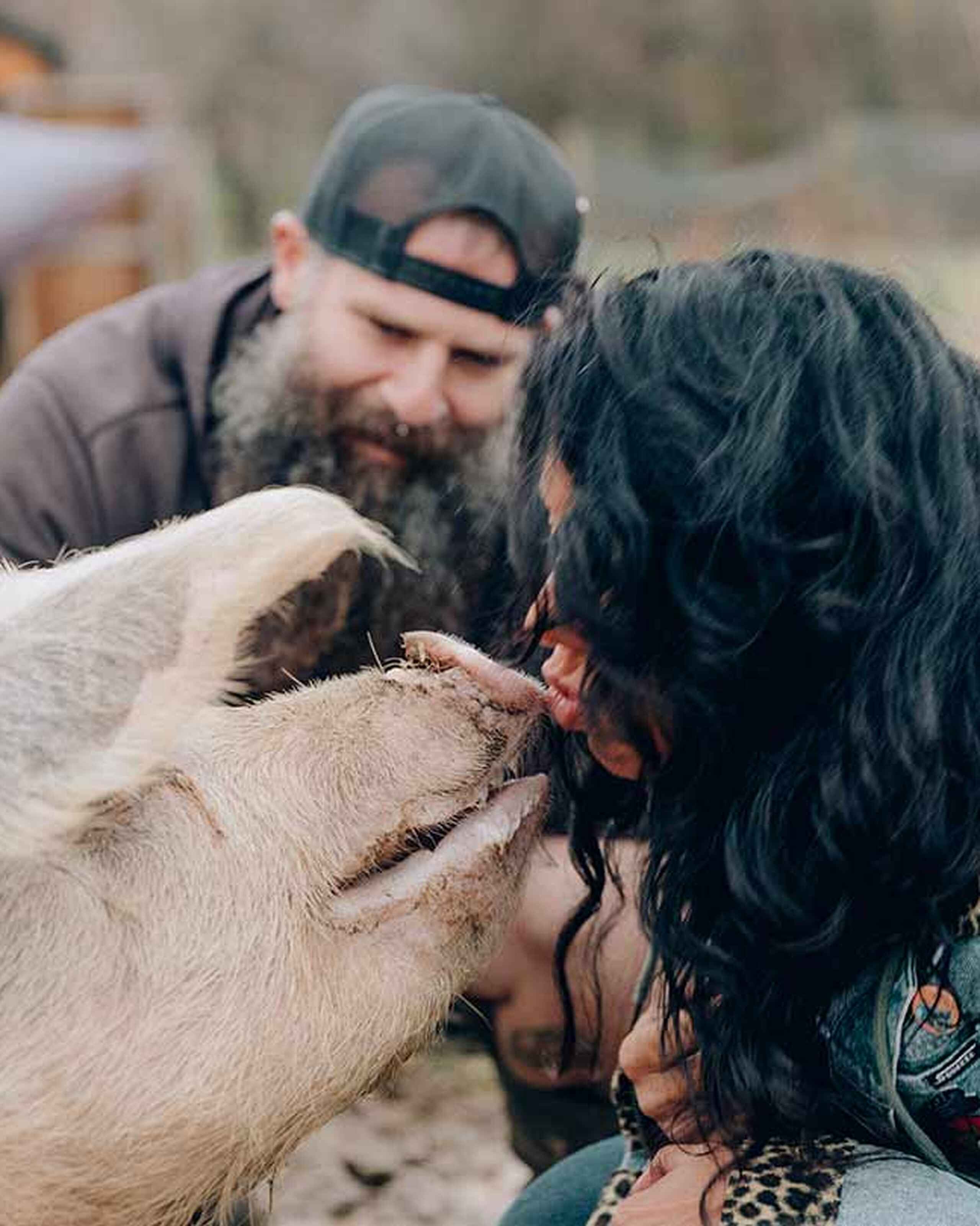 A woman with long dark hair interacts with a white and tan goat while a bearded man in a cap watches nearby on a farm
